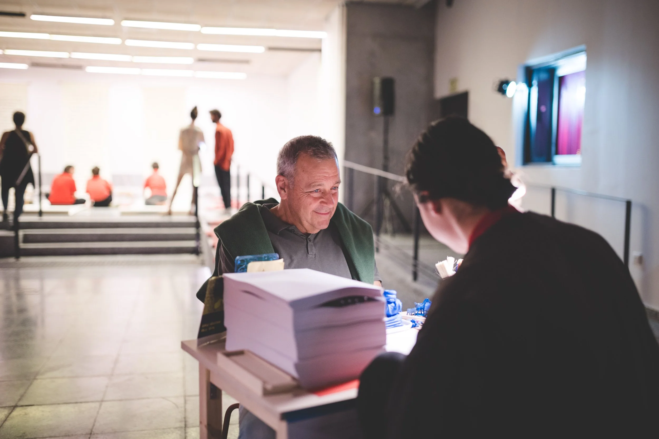 An older man receiving assistance at a registration or information desk inside a modern building, with several people in the background, some sitting on stairs and others walking or standing near a window.