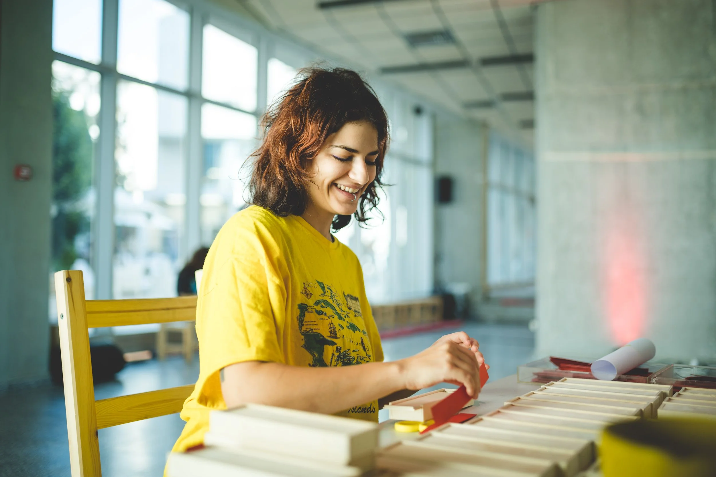 A young woman with curly hair and a yellow t-shirt smiling while working on a craft project at a table with organized craft supplies.