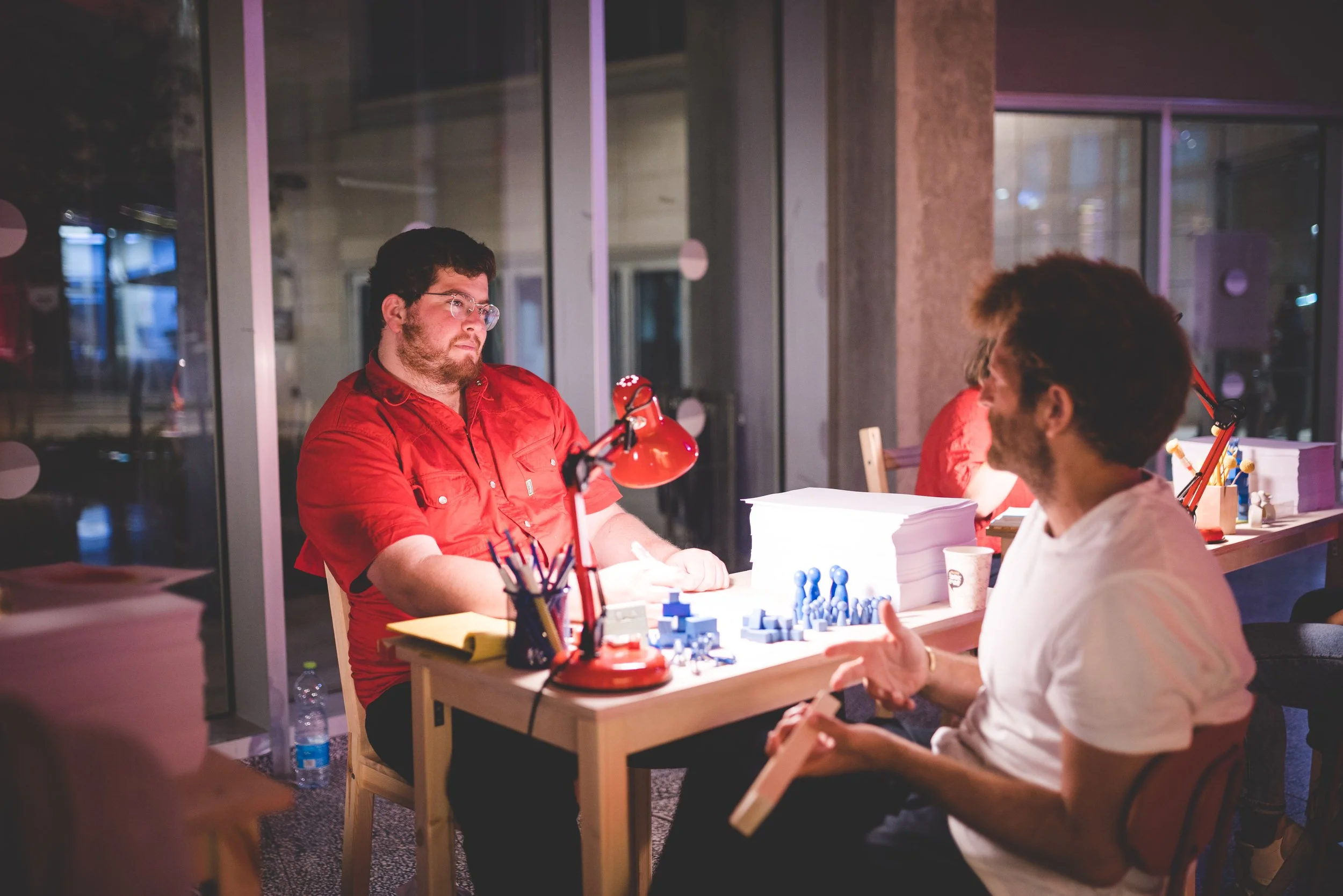 Two men having a conversation at a table in an indoor setting, with stacks of papers and small figurines on the table, and a woman in the background.