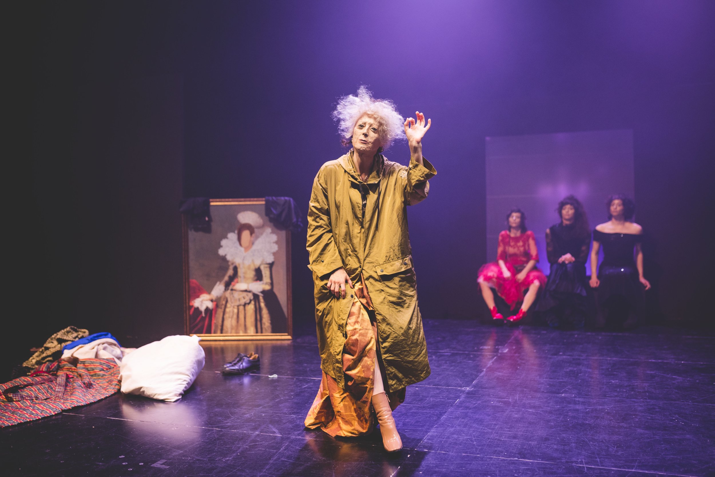 Stage scene with a woman with wild white hair speaking or singing, dressed in a long oversized brown coat and yellow dress, gesturing with her right hand. In the background, three women in colorful dresses sit against a dark purple backdrop. Stage pr