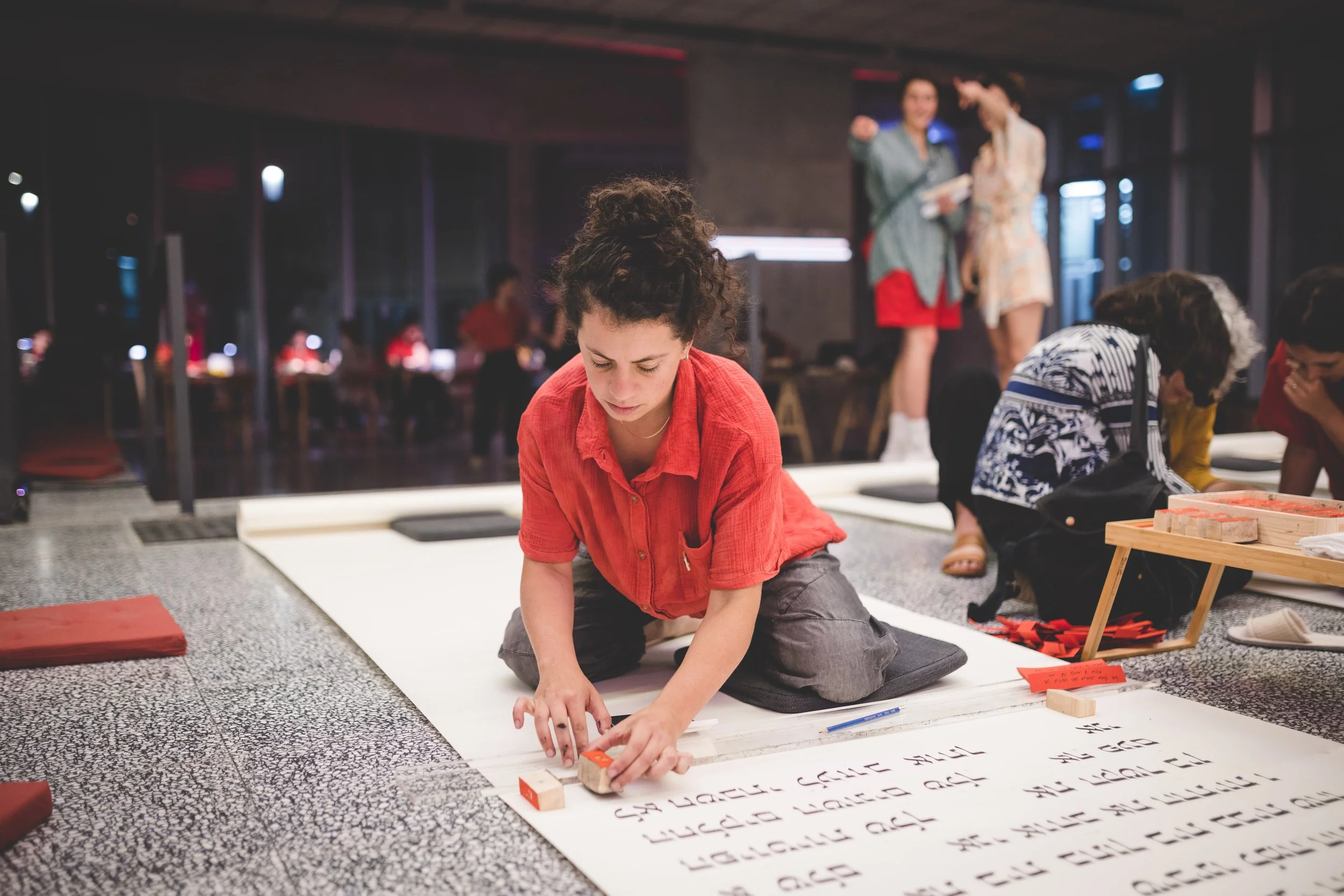 A young woman in a red shirt kneels on the floor, writing on a large white sheet with black marker, in a room with other people engaged in similar activities. Two women are standing and talking in the background.