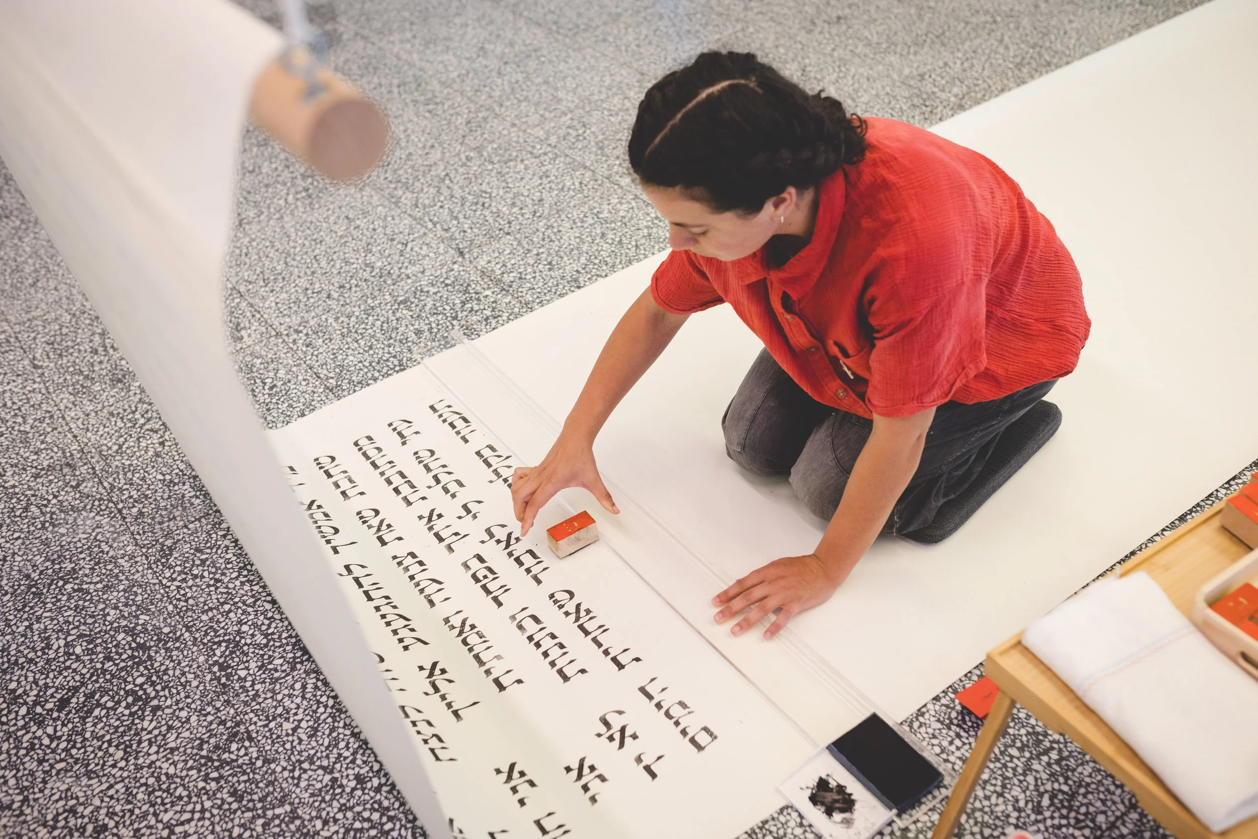 A person in a red shirt kneeling on a mat, using a tool to create a calligraphy piece on white paper, with a smaller table nearby holding additional supplies.