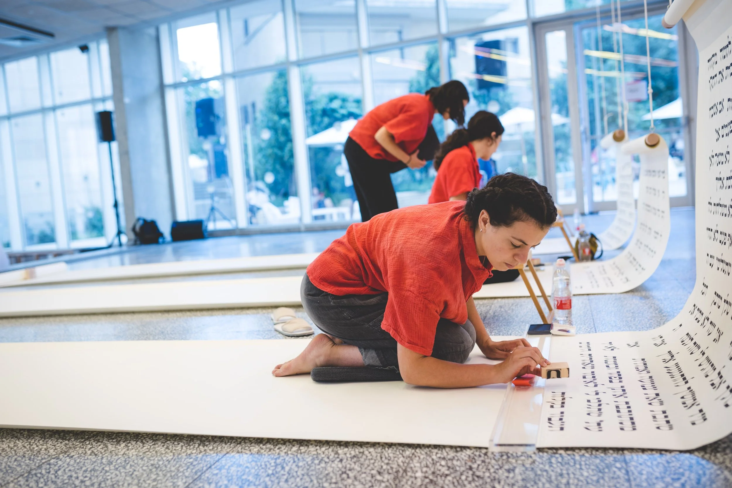 Women in red shirts working on a large scroll with text in a modern, glass-walled space.