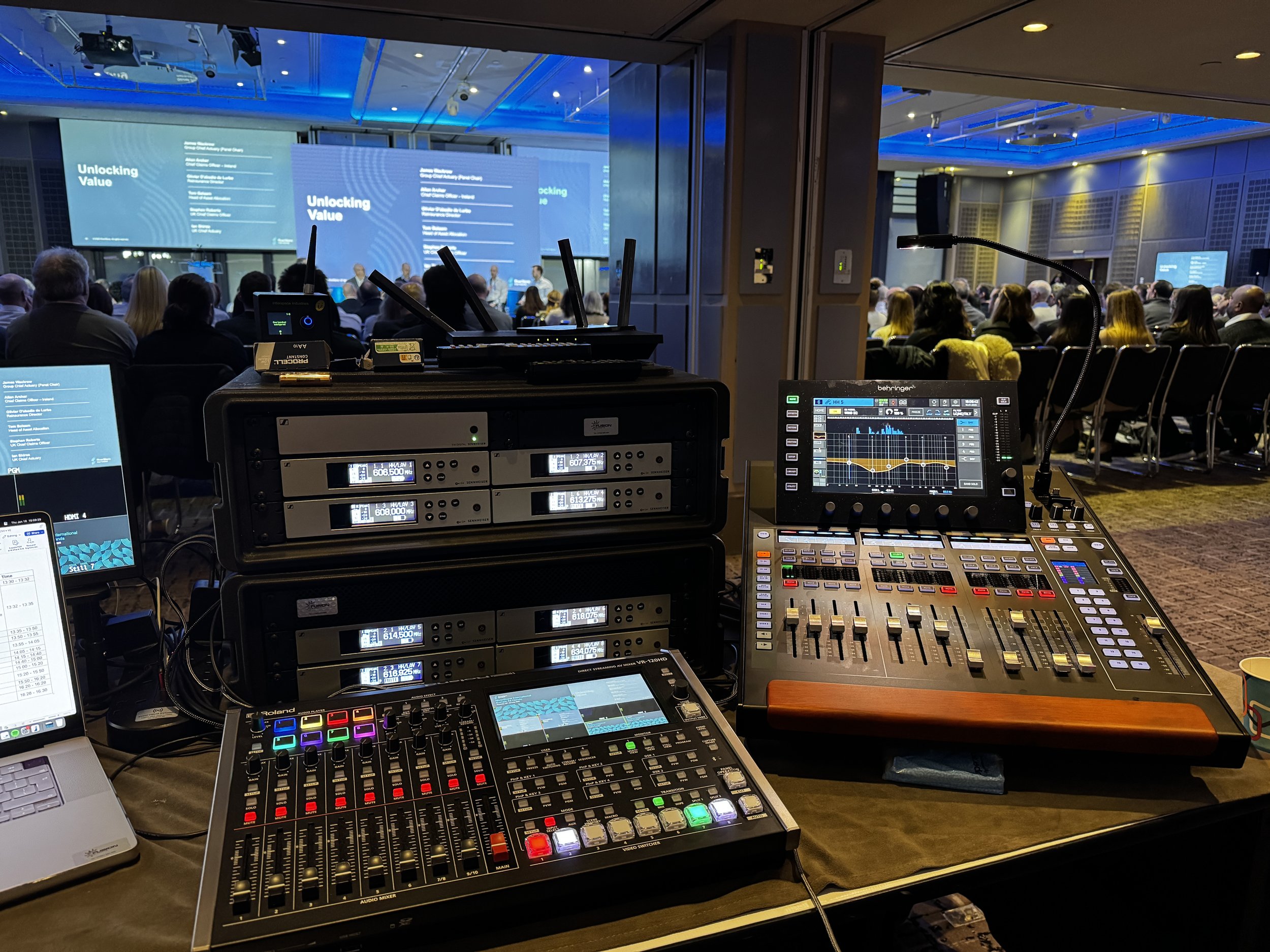 Conference or seminar room with a stage, large screens displaying 'Unlocking Value', and an audience seated facing the stage. Audio-visual equipment and sound mixing consoles are in the foreground.