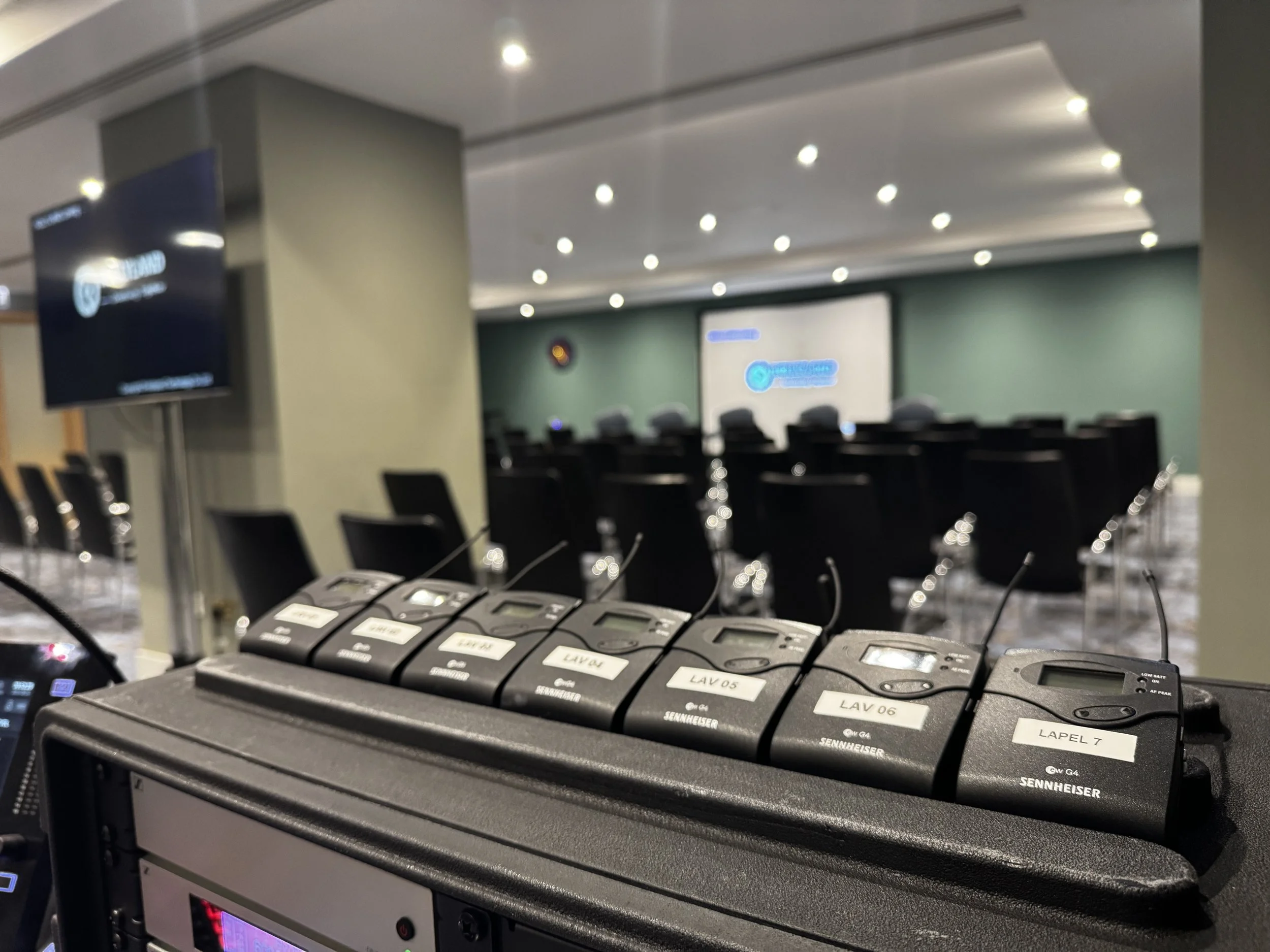 Wireless microphone transmitters lined up on a rack in an auditorium or conference room with chairs and a projector screen in the background.