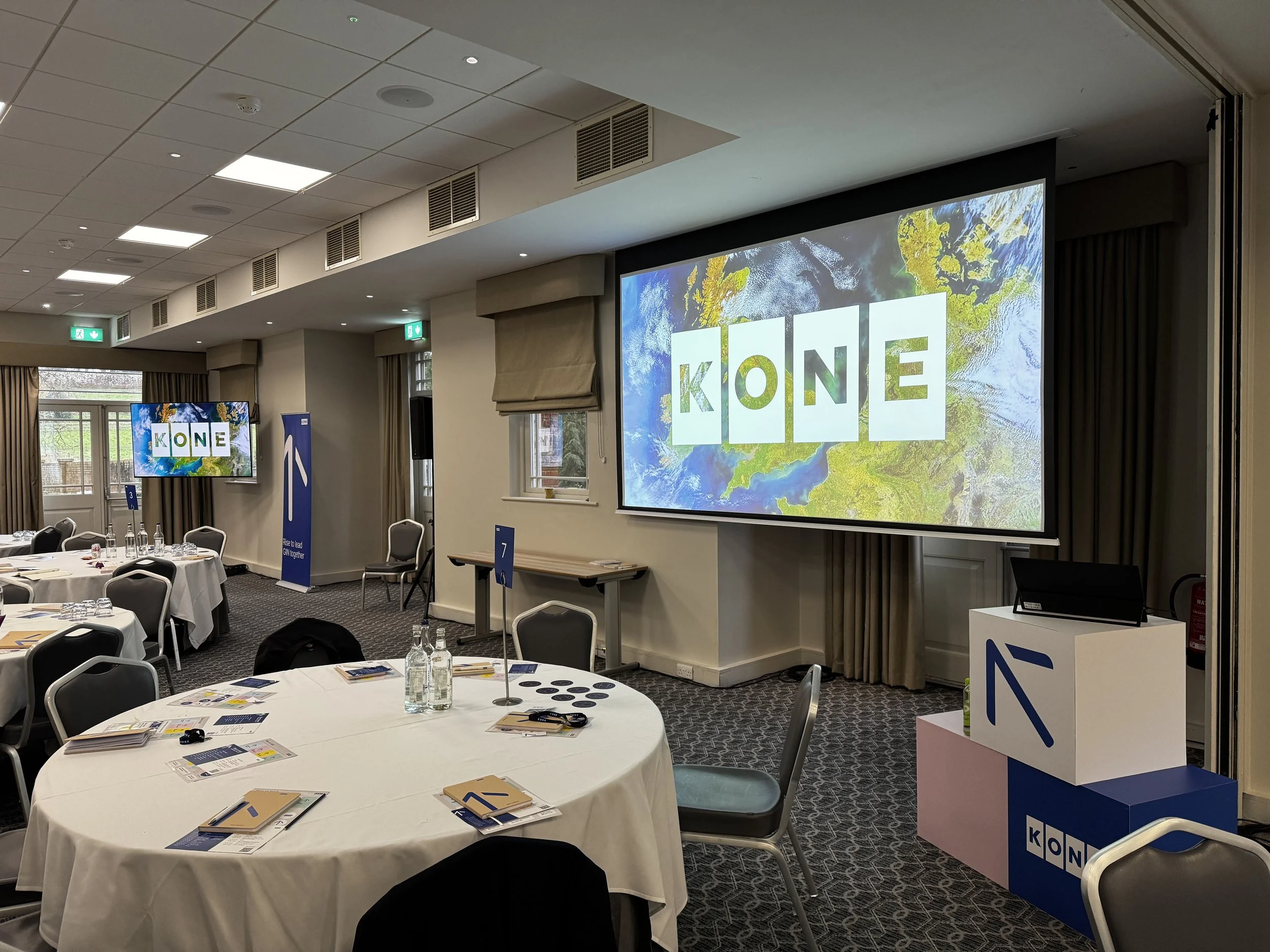 Conference room with round tables set for a meeting, displaying brochures and water bottles. Large screen at the front shows the KONE logo over a map background. Additional smaller screens and signage with the KONE branding are visible in the room.
