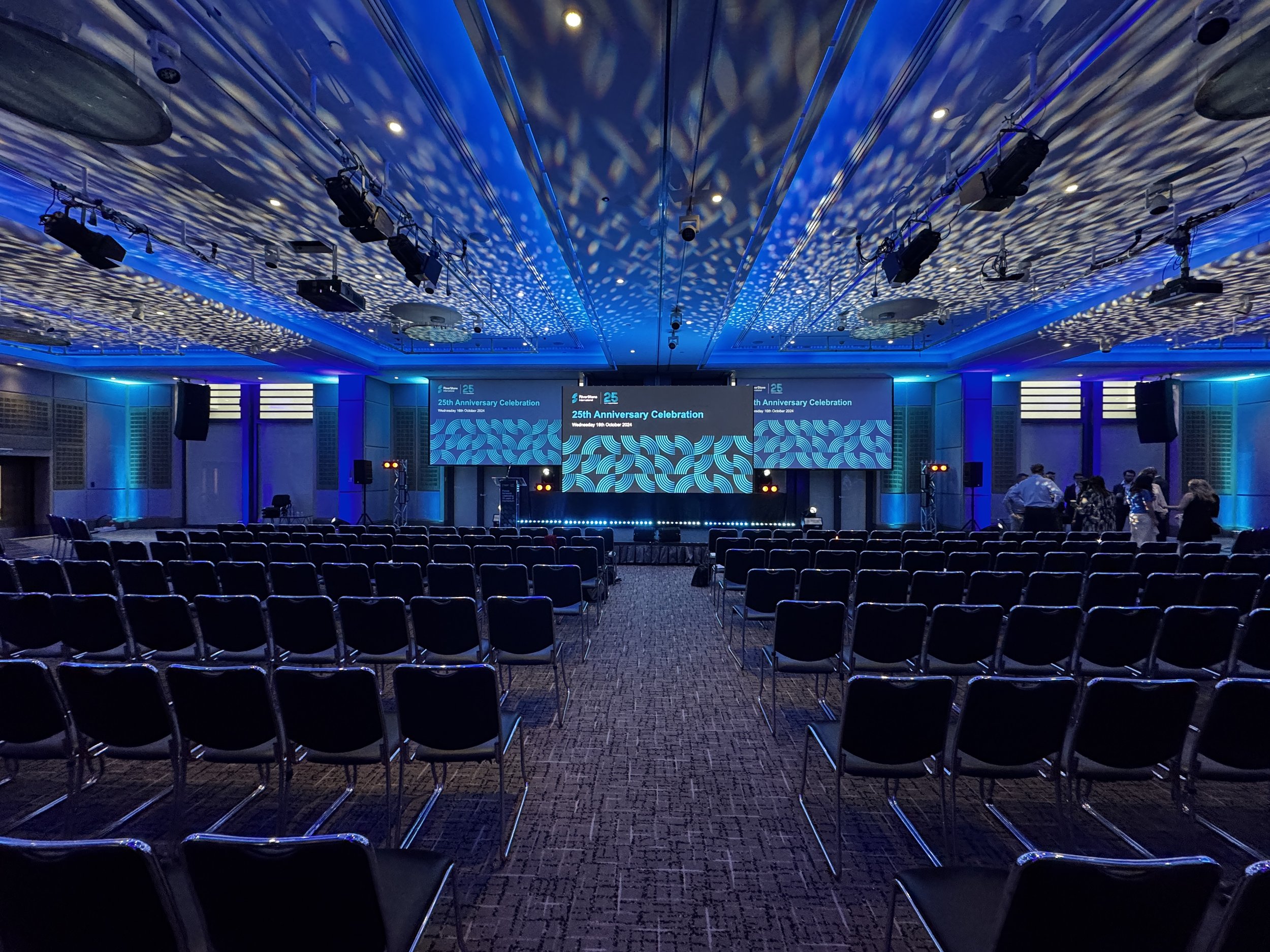 An empty conference room set up for a 25th anniversary celebration, with rows of black chairs facing a stage with three large screens displaying event information and blue lighting creating a patterned ceiling effect.