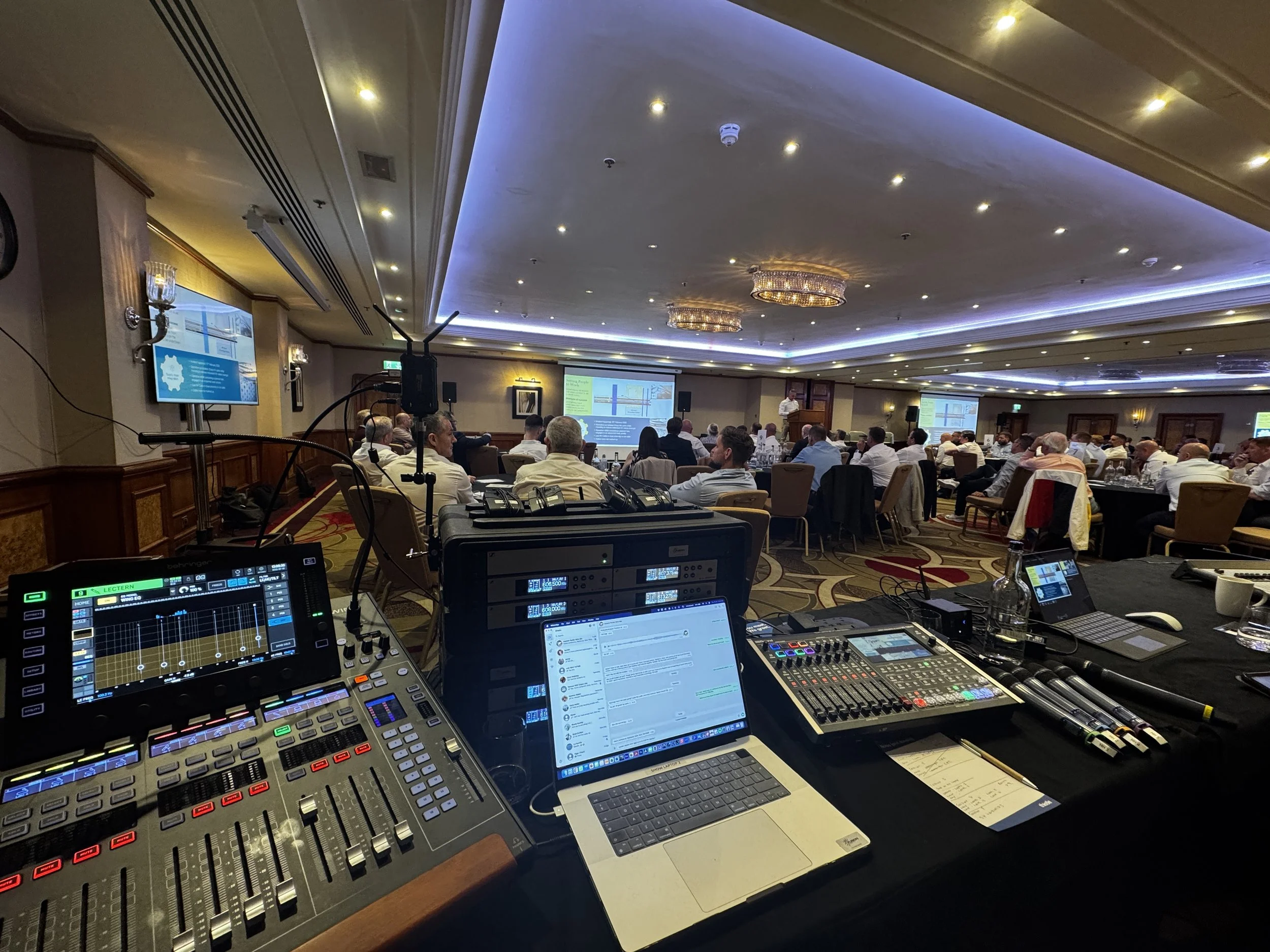 Conference room with audience watching a speaker at a podium, multiple screens displaying presentation, viewed from audio and lighting equipment station in foreground.