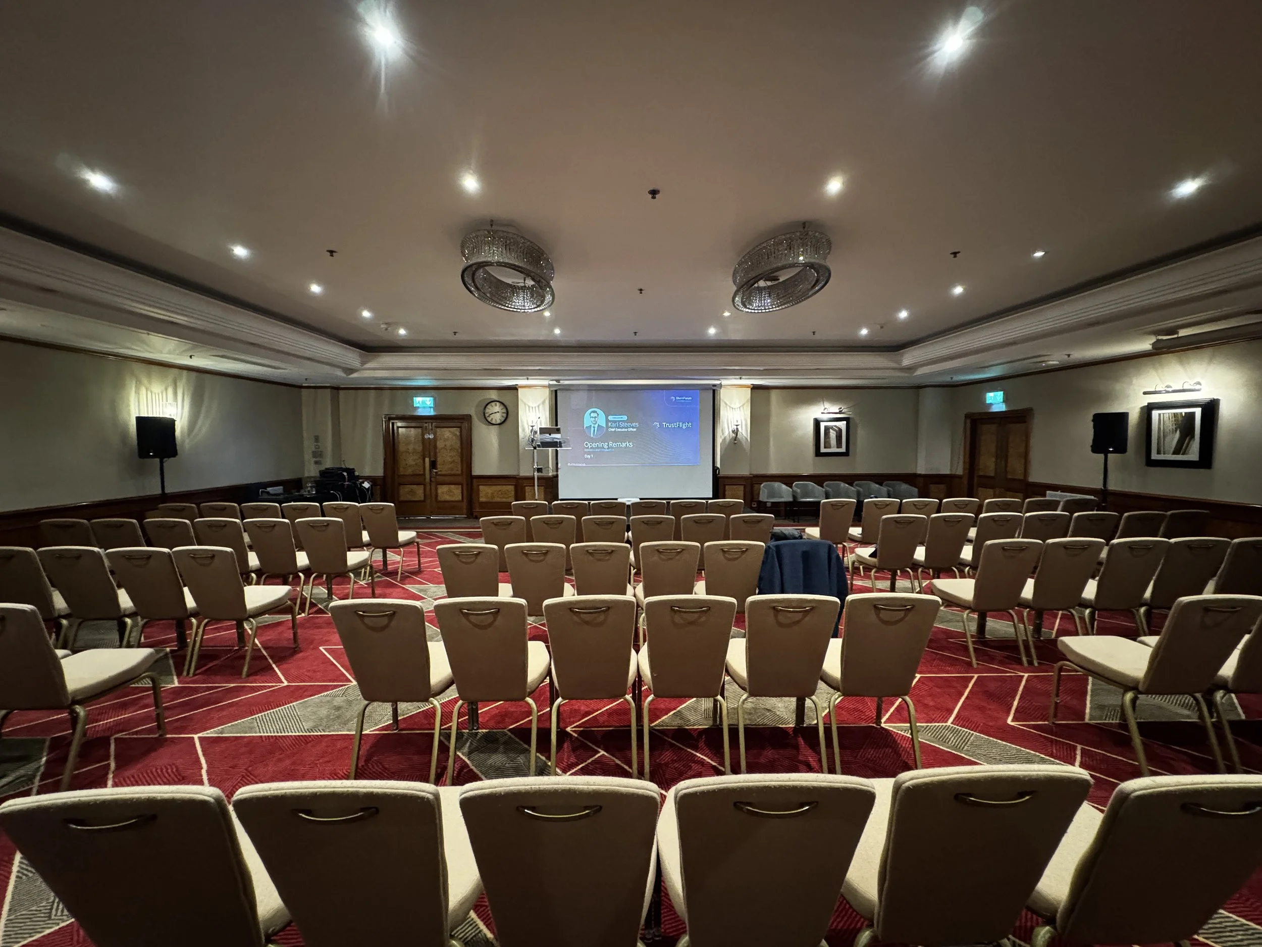 Empty conference room with rows of beige chairs facing a stage with a presentation screen, two chandeliers, and a red patterned carpet.