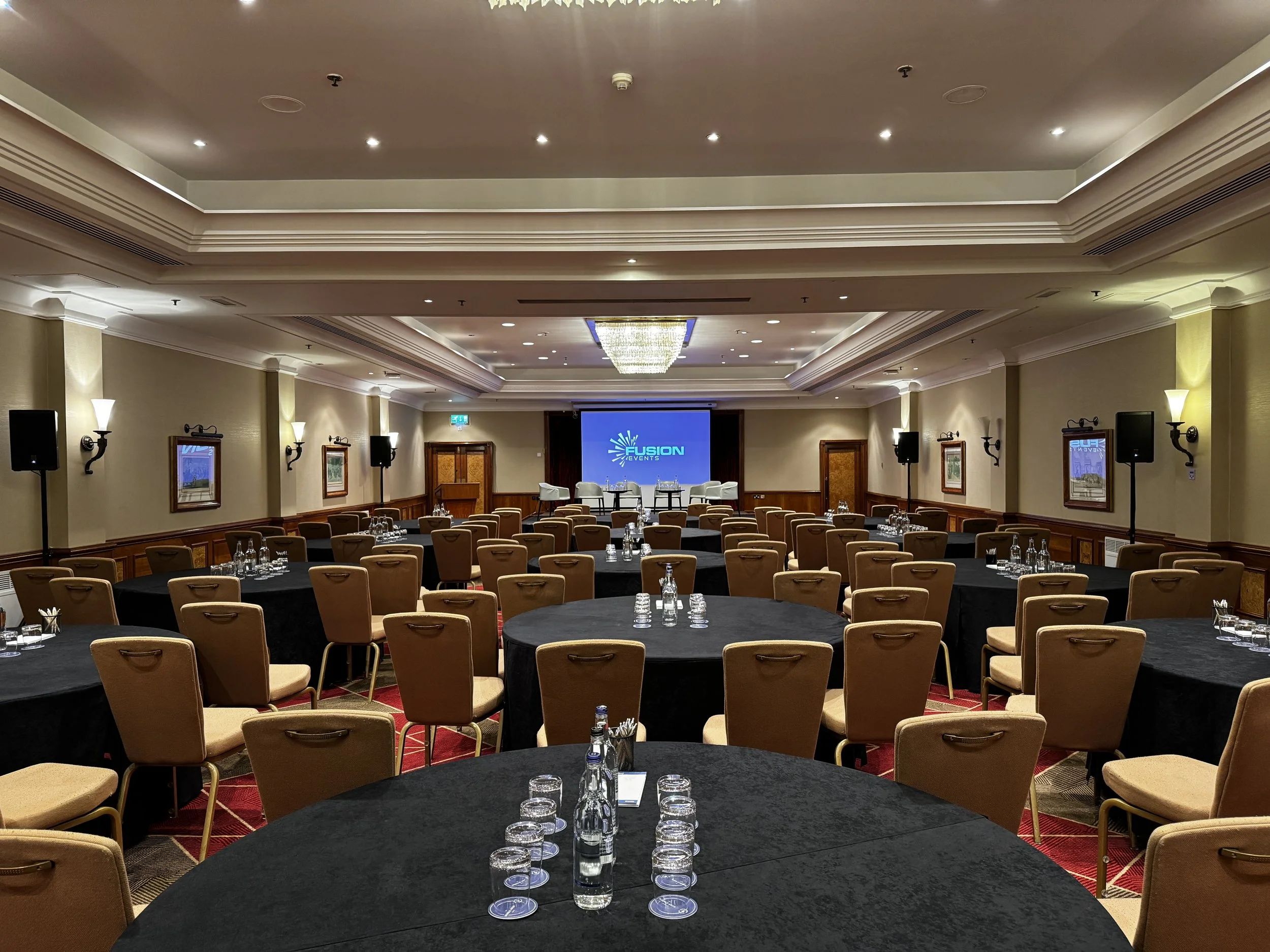 Conference room set up with round tables covered in black tablecloths, beige chairs, and a stage with a large screen displaying the Fusion logo at the front.