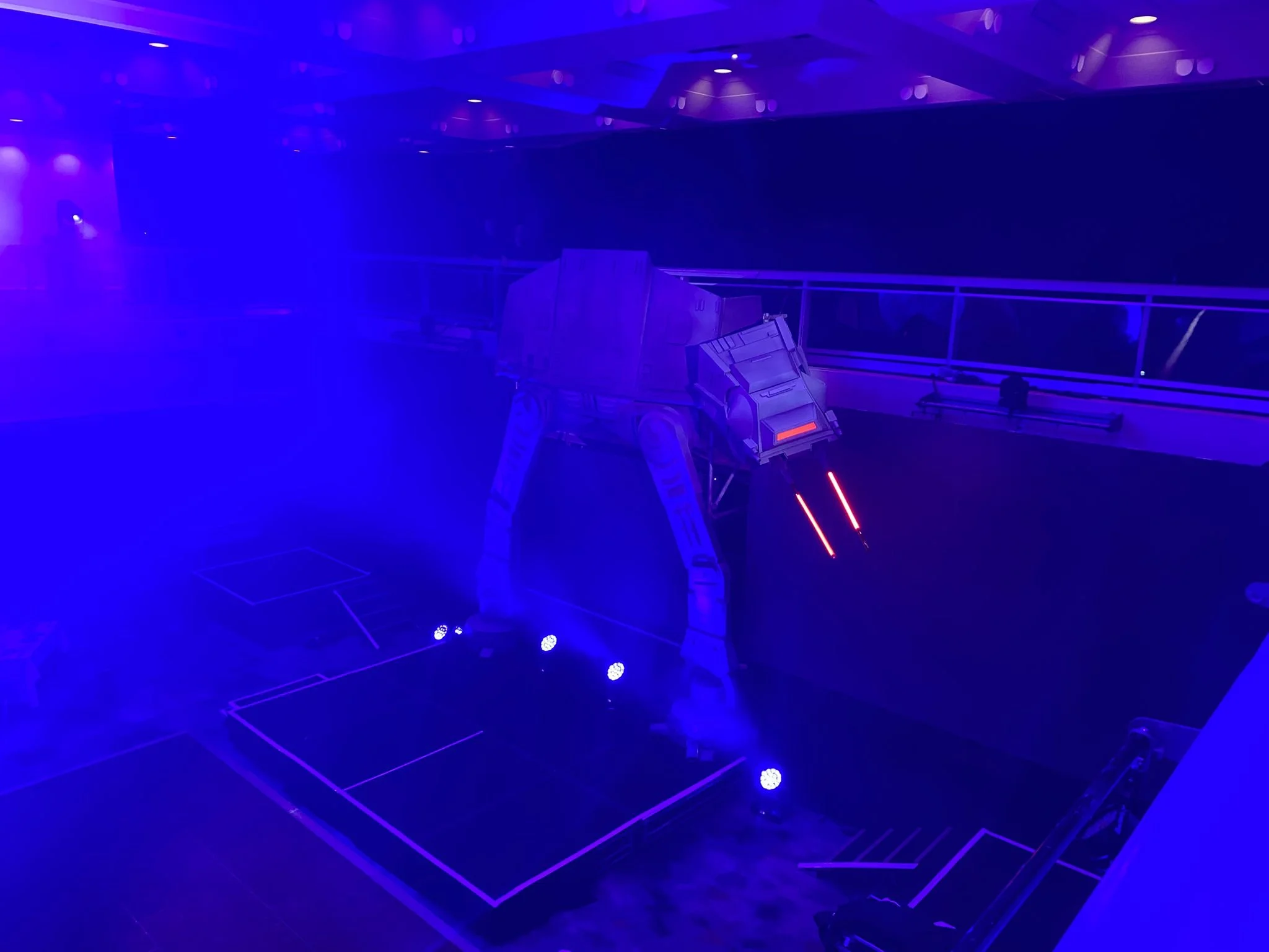 An indoor scene with a large, metallic AT-AT Walker from Star Wars, illuminated in blue with some red accents, standing on a black floor with white outline markings, in a dimly lit environment.