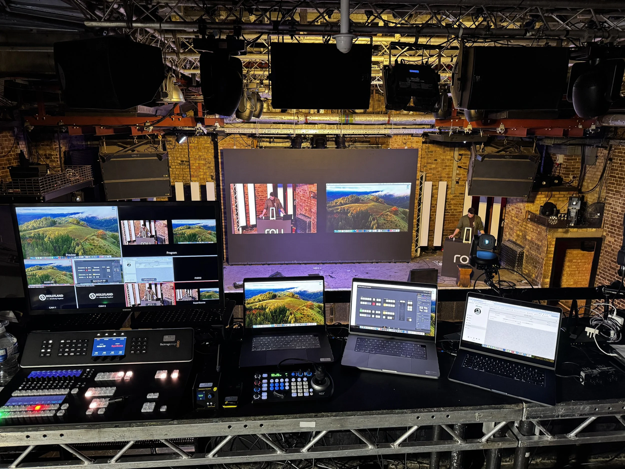 Stage setup with large screens, control panels, laptops, and a person working at a DJ booth in a brick-walled venue.