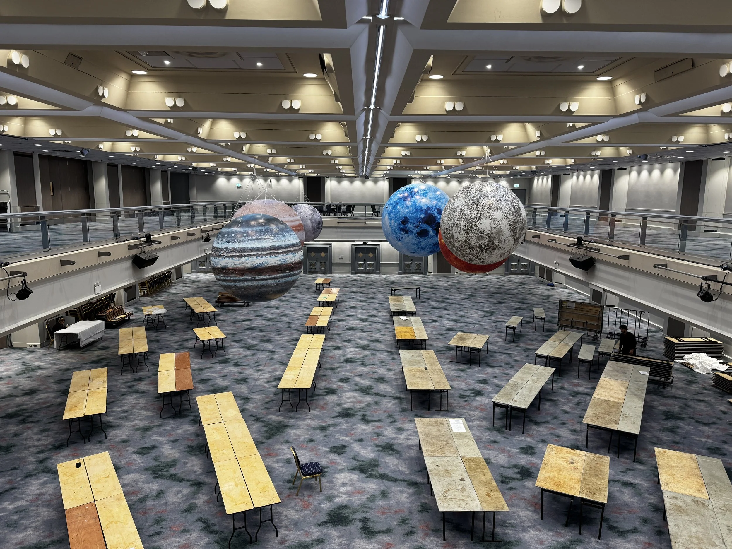 A large, empty conference room with a high ceiling, decorated with planets hanging from the ceiling, and multiple rows of tables and chairs arranged on a patterned carpet.