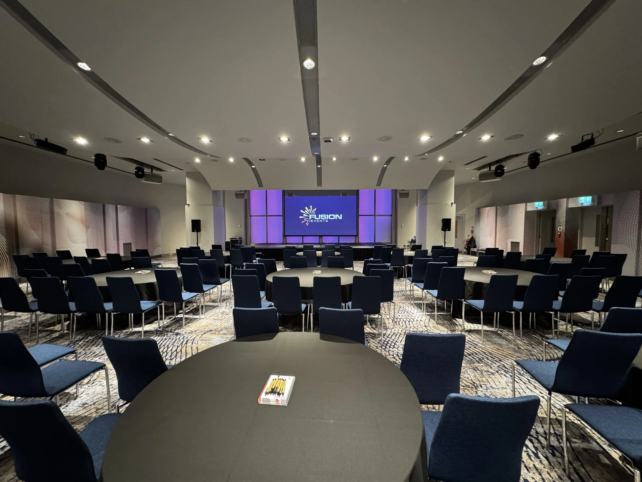 Empty conference room with round tables and blue chairs facing a stage with a large screen displaying Fusion Events logo.