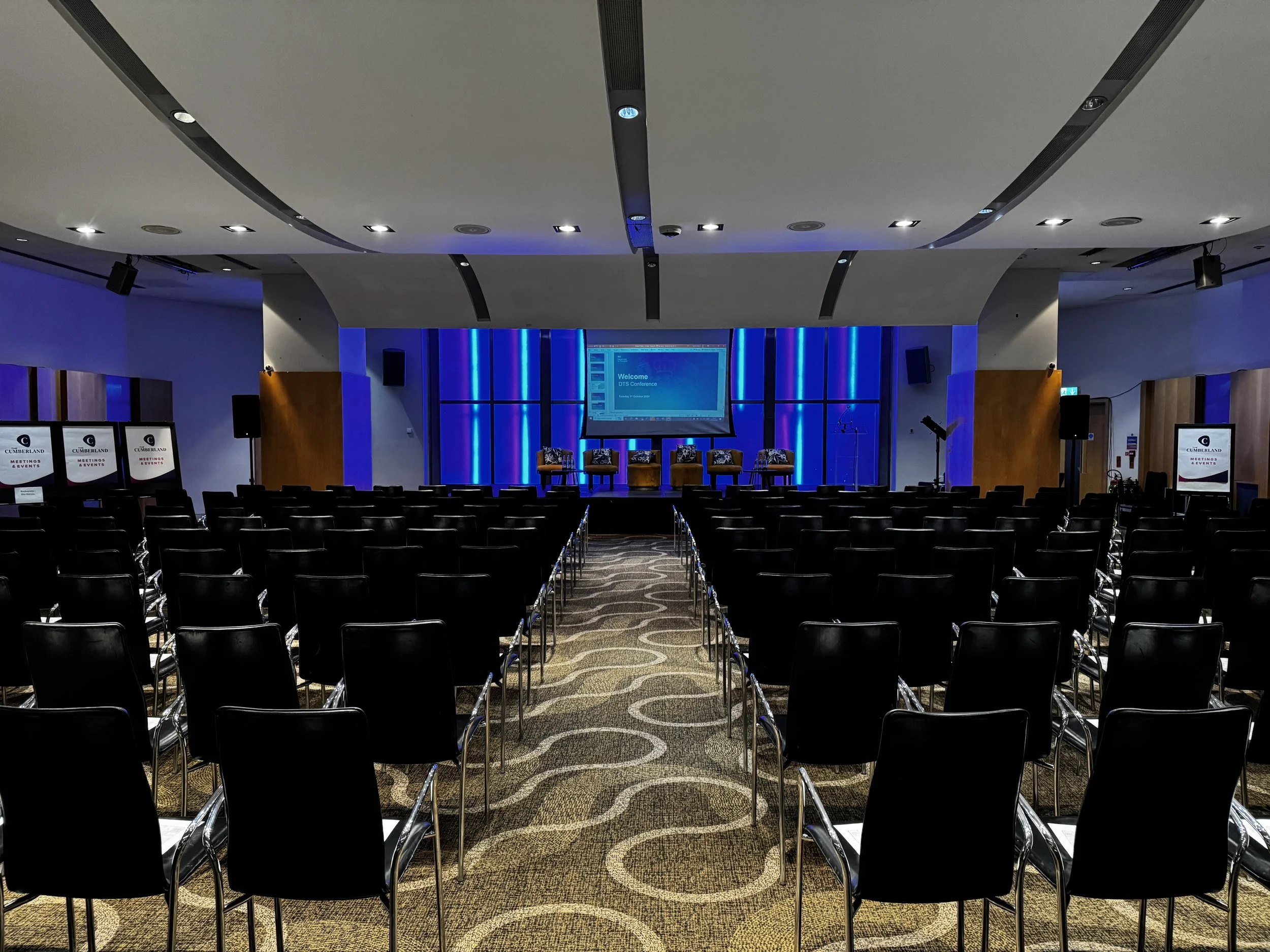 Empty conference room with rows of black chairs facing a stage with a screen and four chairs.