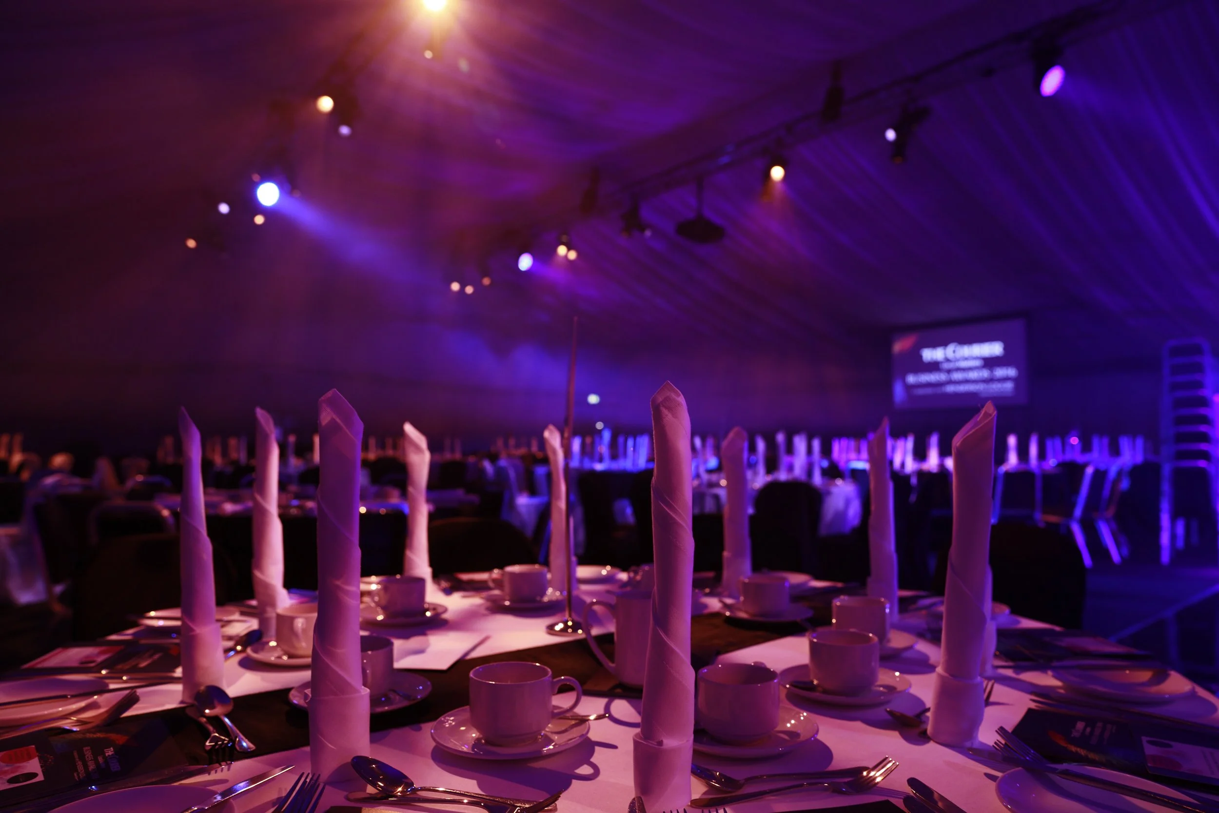 Tables set with white napkins, cups, saucers, and silverware in a dimly lit event hall with purple and orange lighting, a large screen in the background, and a crowd of people seated.