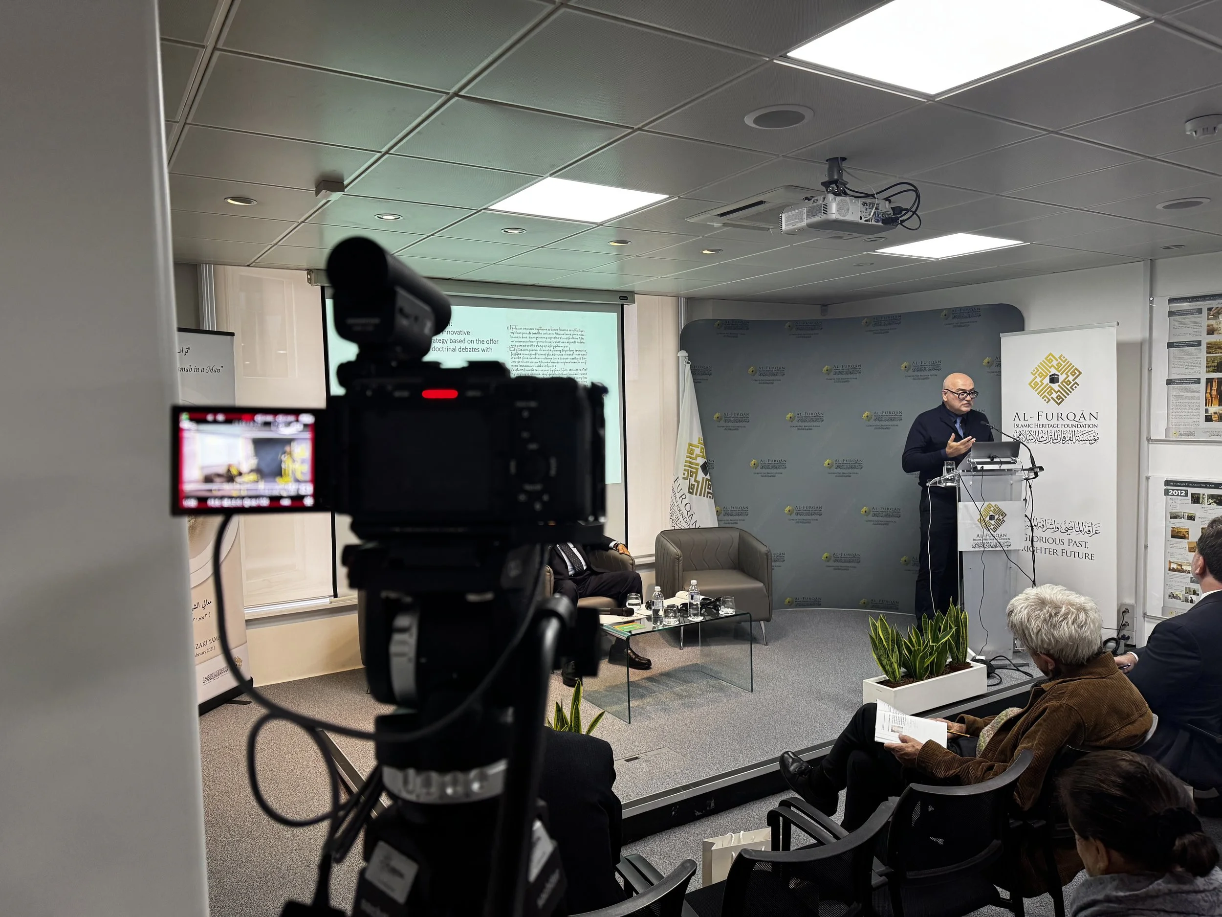 A man in a dark suit and glasses giving a presentation at a podium in a conference room with a banner that reads 'Al Furqan' with an emblem. Attendees are seated, some reading documents, and a camera is recording the event.