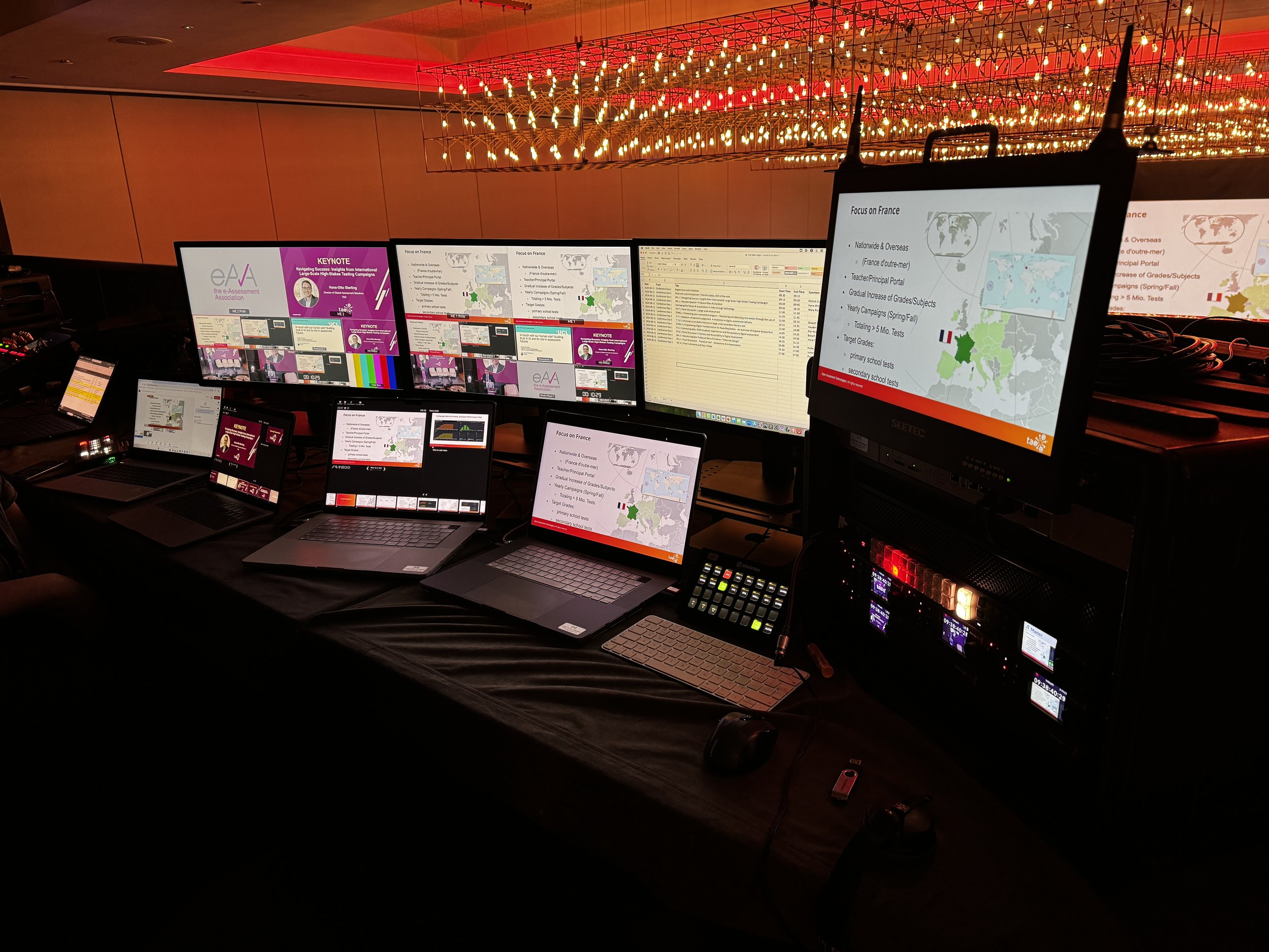 Multiple computer monitors and laptops on a table displaying slides and data related to a conference or presentation, with a decorative ceiling light fixture above.