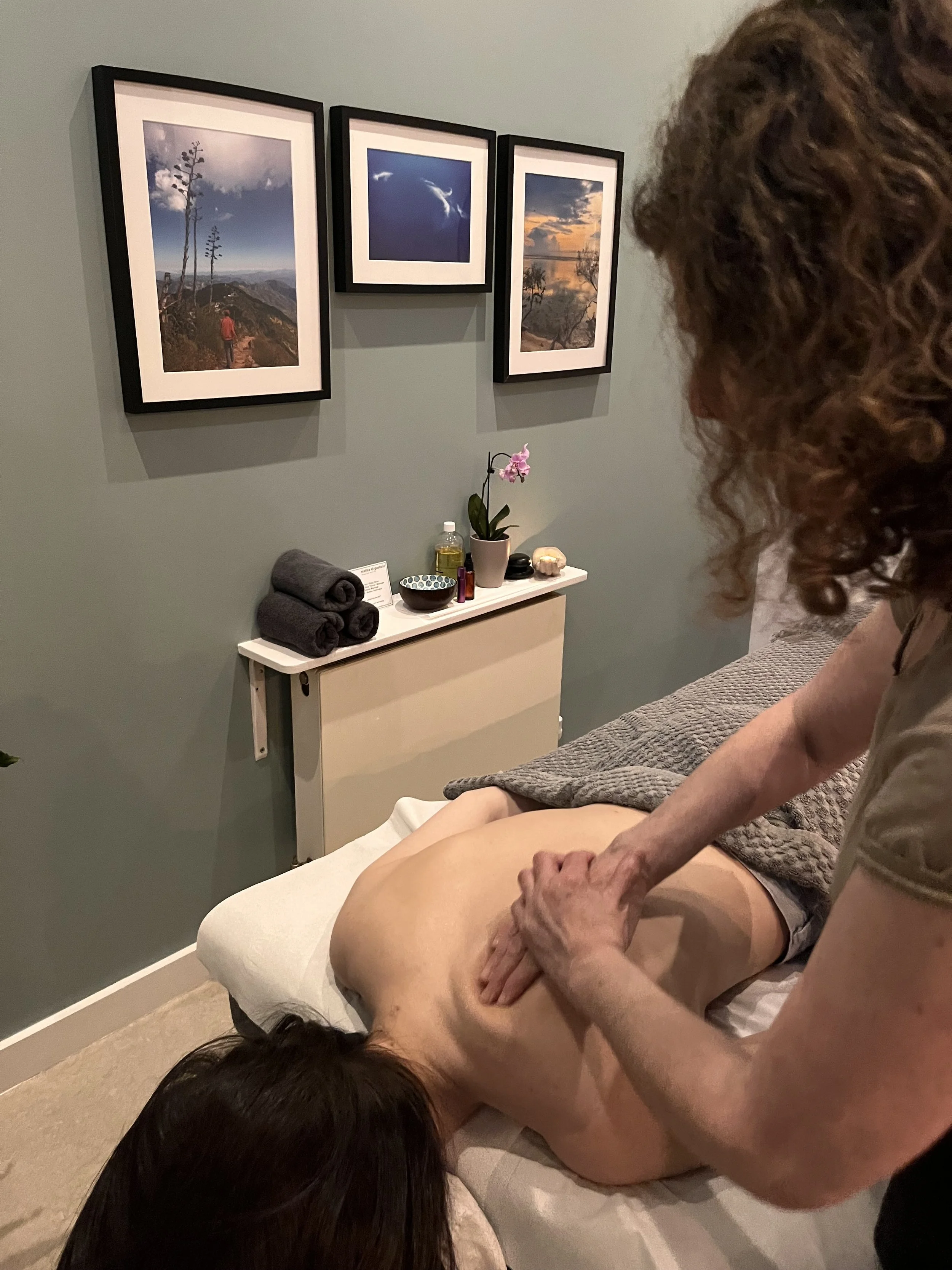 A person receiving a massage in a spa room with framed landscape photos on the wall and a table with towels, a potted orchid, and massage supplies.