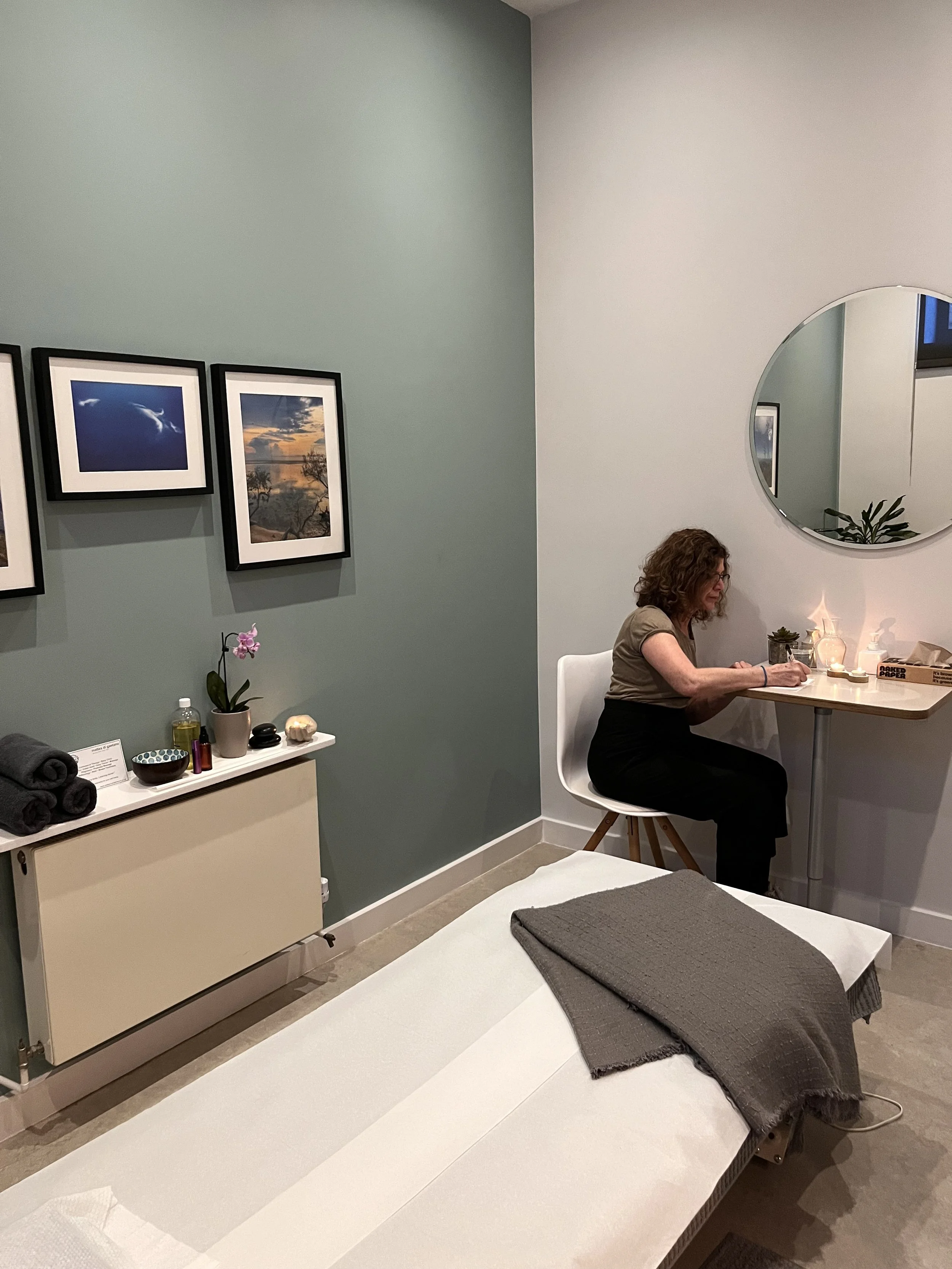 A woman sitting at a small wooden table with candles and a plant, writing, in a wellness or massage room with a massage table covered in white sheets and a gray blanket.