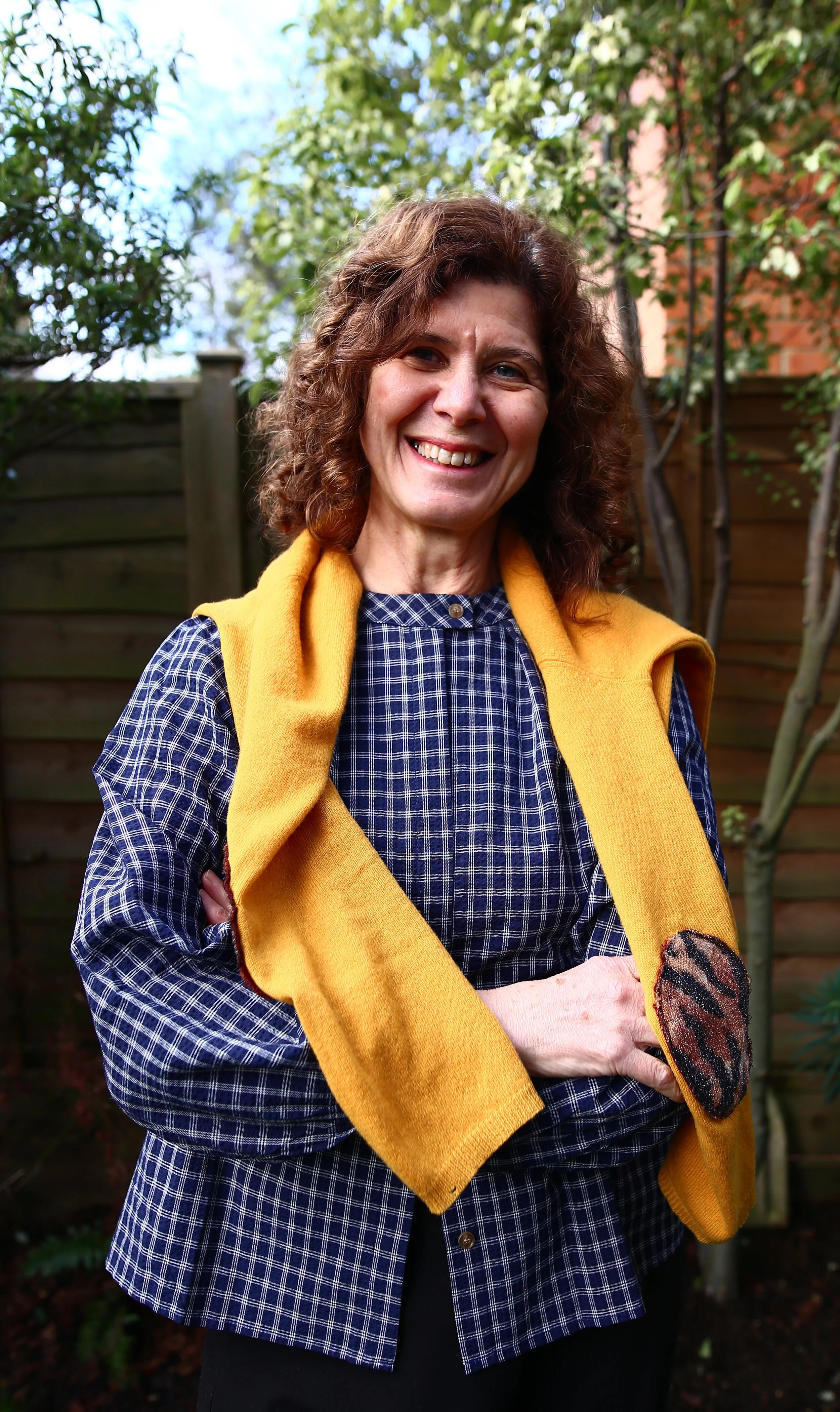 A woman with curly brown hair and fair skin stands outdoors in front of a wooden fence, smiling at the camera. She wears a blue plaid dress and has a yellow sweater draped over her shoulders with a butterfly patch on the sleeve.