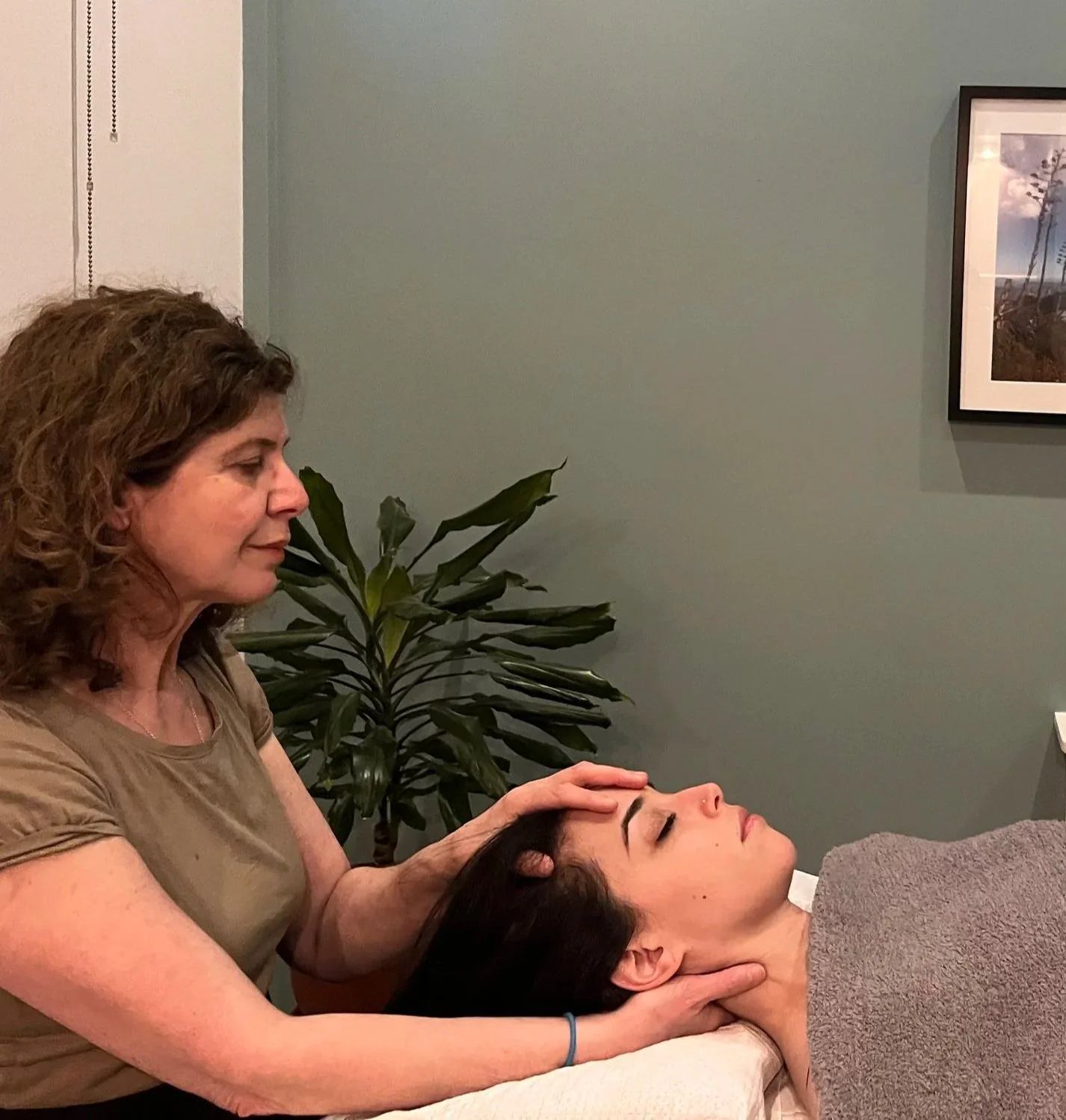 A woman giving a facial massage or therapy to a woman lying on a massage table, with green plants and framed art on the wall in the background.