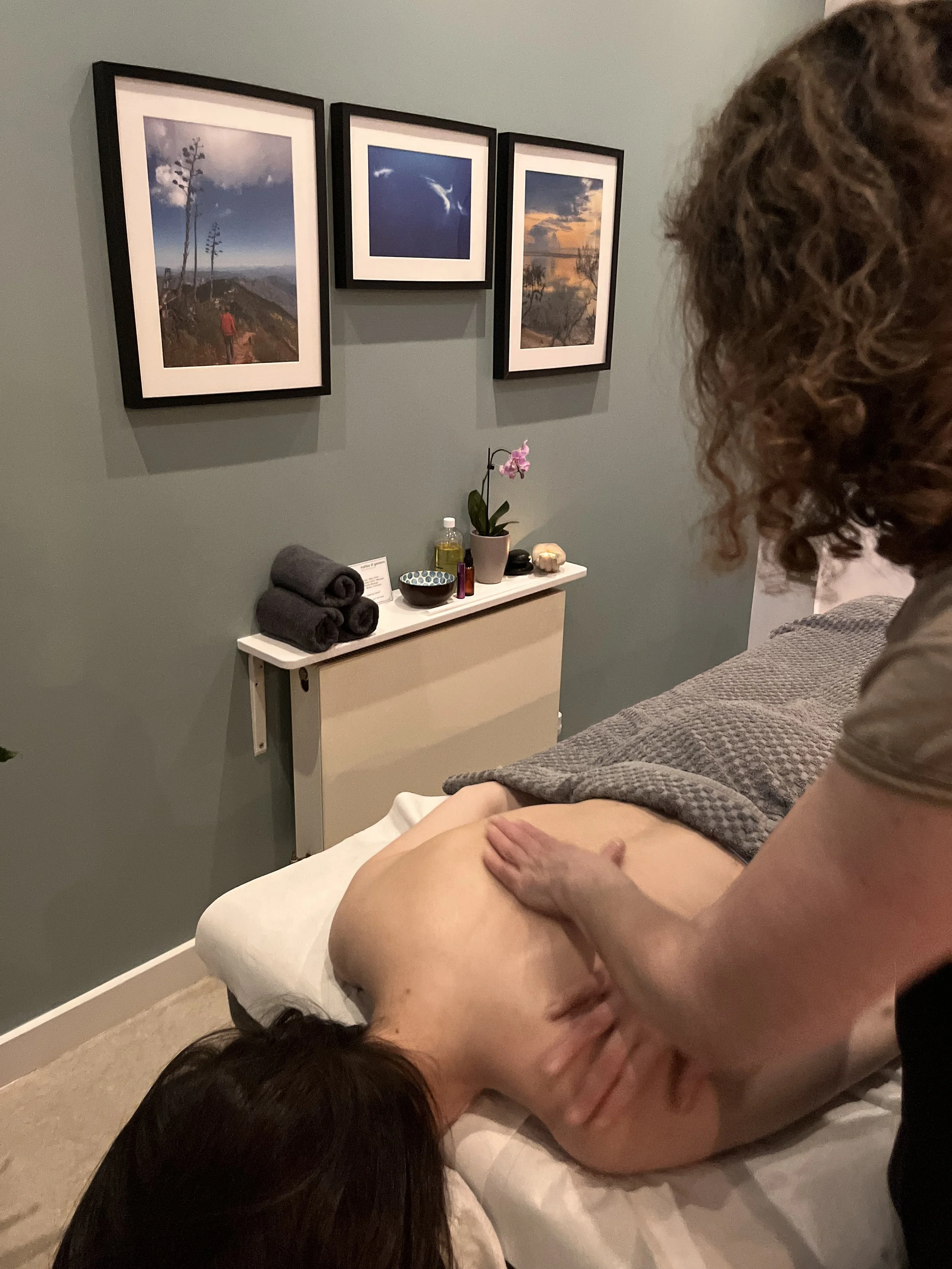 Person receiving a massage in a wellness or spa room with framed landscape photos on the wall and a table with rolled towels, a potted plant, and massage oils.