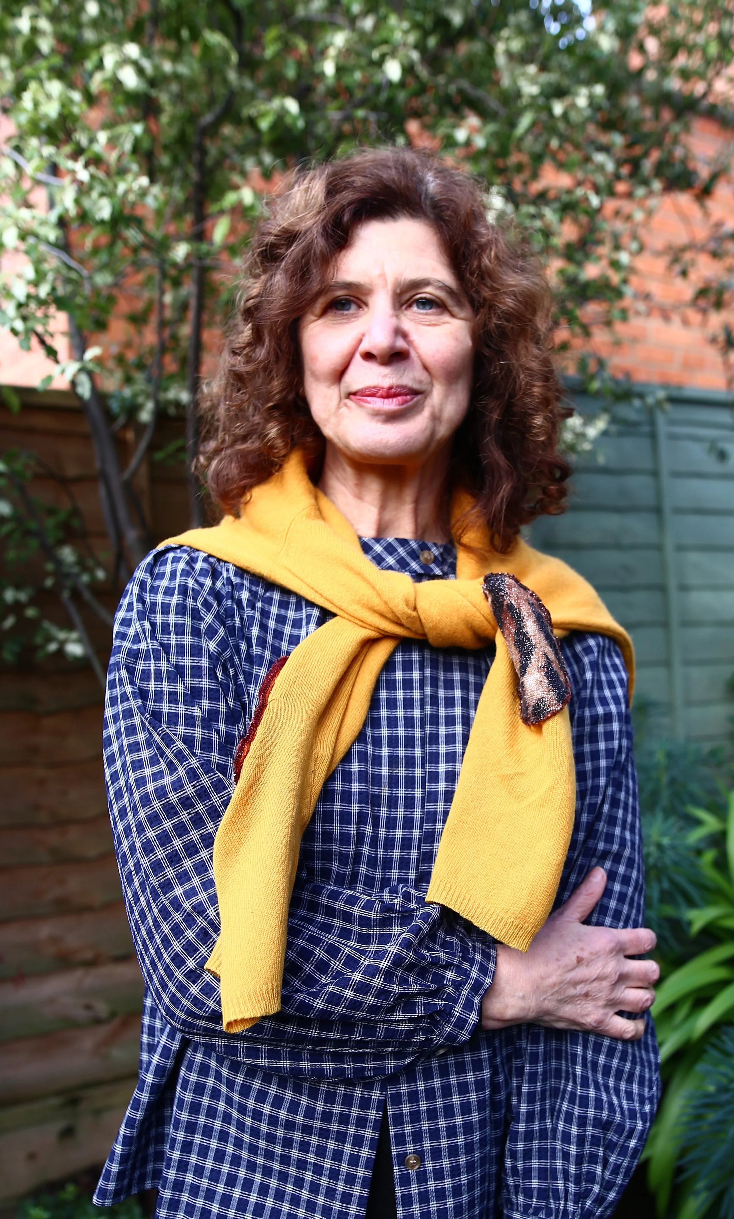 A middle-aged woman with curly brown hair and blue eyes standing outdoors in a garden with a brick wall and green plants in the background. She is wearing a blue plaid dress with a yellow sweater draped over her shoulders.