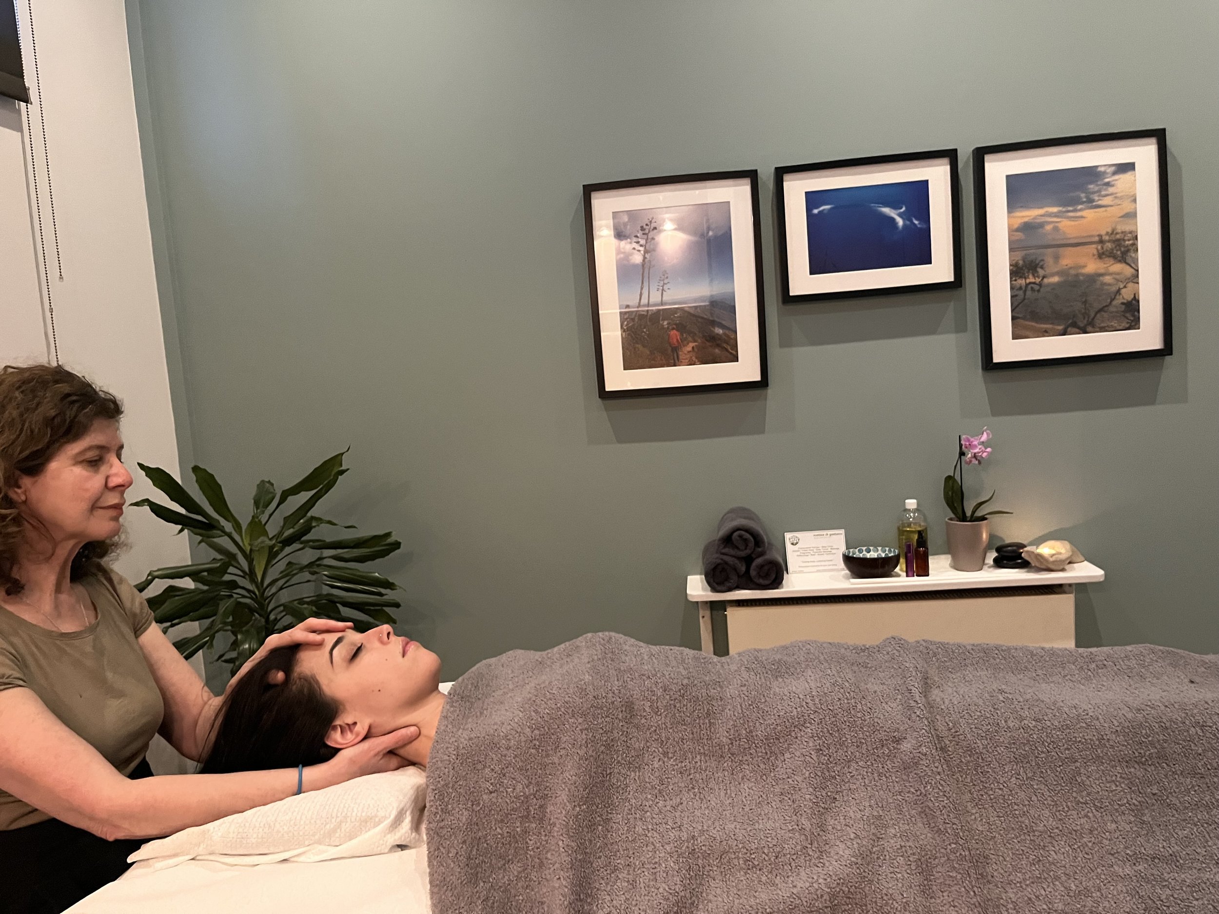 A woman receives a facial massage from a person lying on a treatment table, with calm and relaxed expressions in a spa or massage room decorated with pictures of nature and flowers.