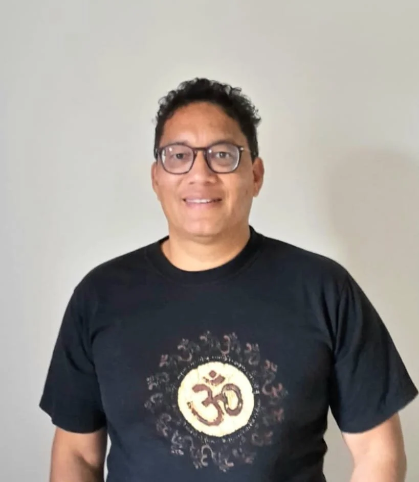 Homem sorrindo, usando óculos e camiseta preta com símbolo Om dourado no centro, parado contra parede clara. 