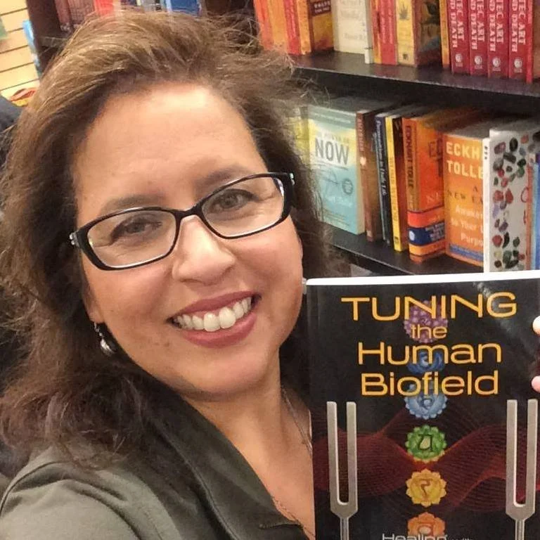Mulher sorridente com óculos segurando um livro intitulado "Tuning the Human Biofield" em uma livraria, com prateleiras cheias de livros ao fundo.