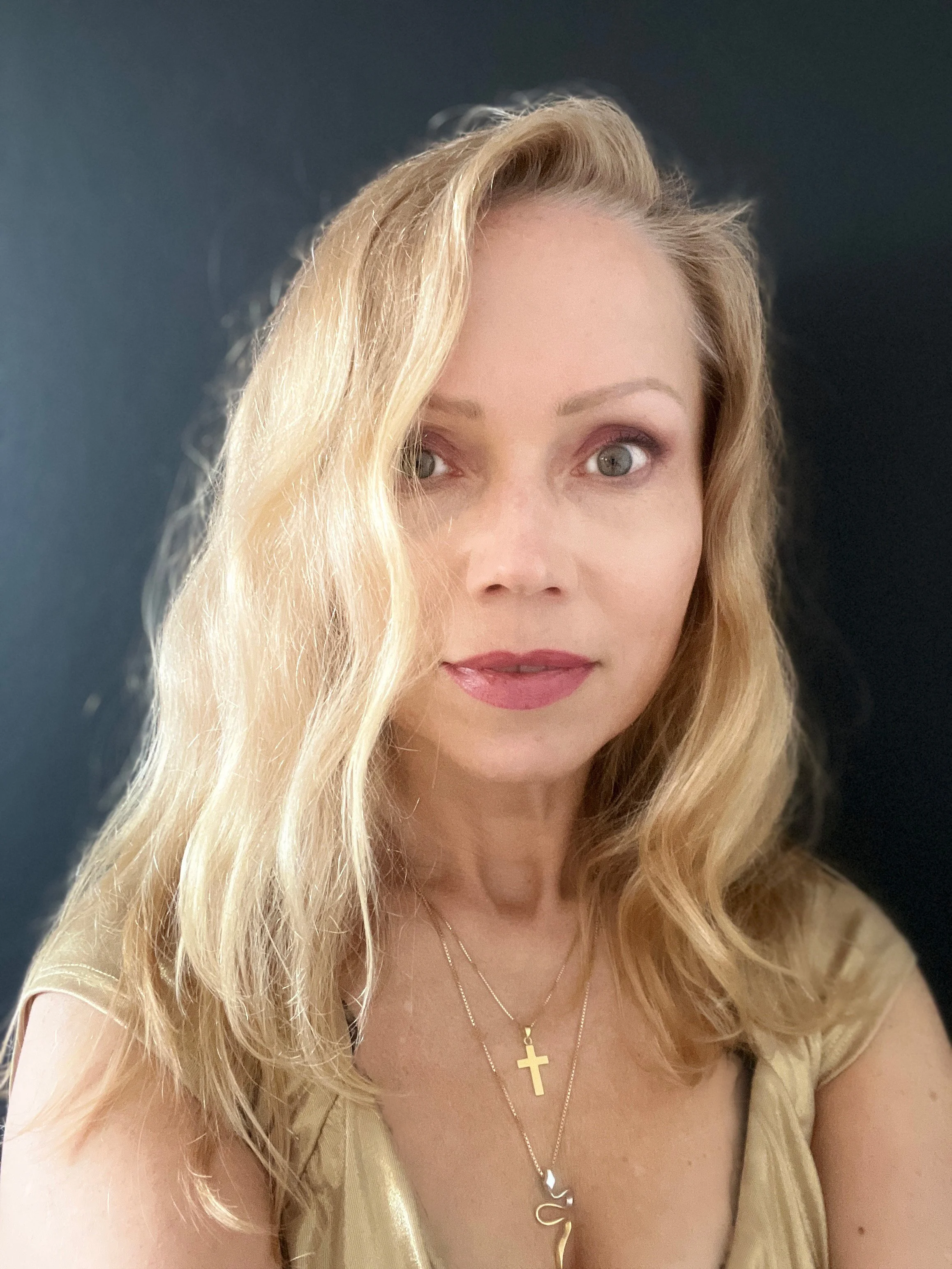 Mulher com cabelos loiros, olhos claros, usando maquiagem, vestido dourado e colares com um crucifixo e uma corrente com um pingente de letra S, contra fundo escuro.