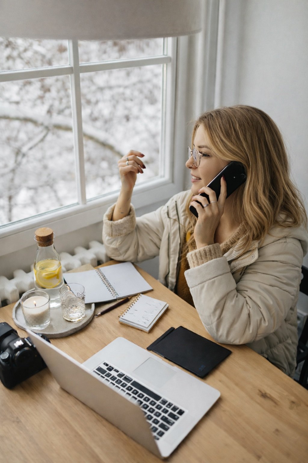 Trauma informed therapist in Hopkins, Minnesota with glasses and red hair sitting at a wooden table near a window, talking on the phone, with a laptop, notebooks, a camera, a glass of water, a candle, and a pitcher of lemon water on the table.