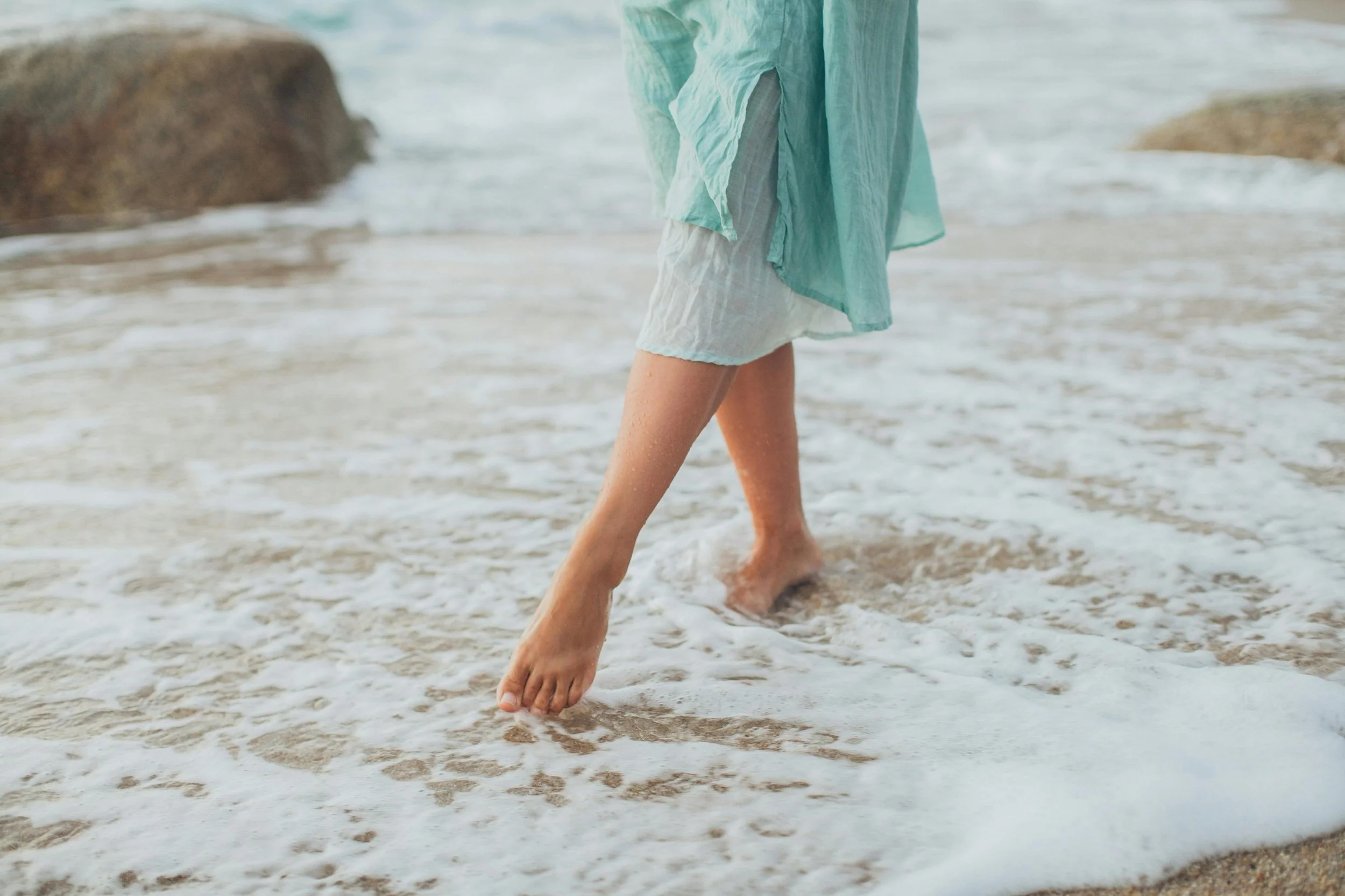 Person walking barefoot along the shoreline, with waves washing over their feet, wearing a light green, flowy cover-up and white shorts.