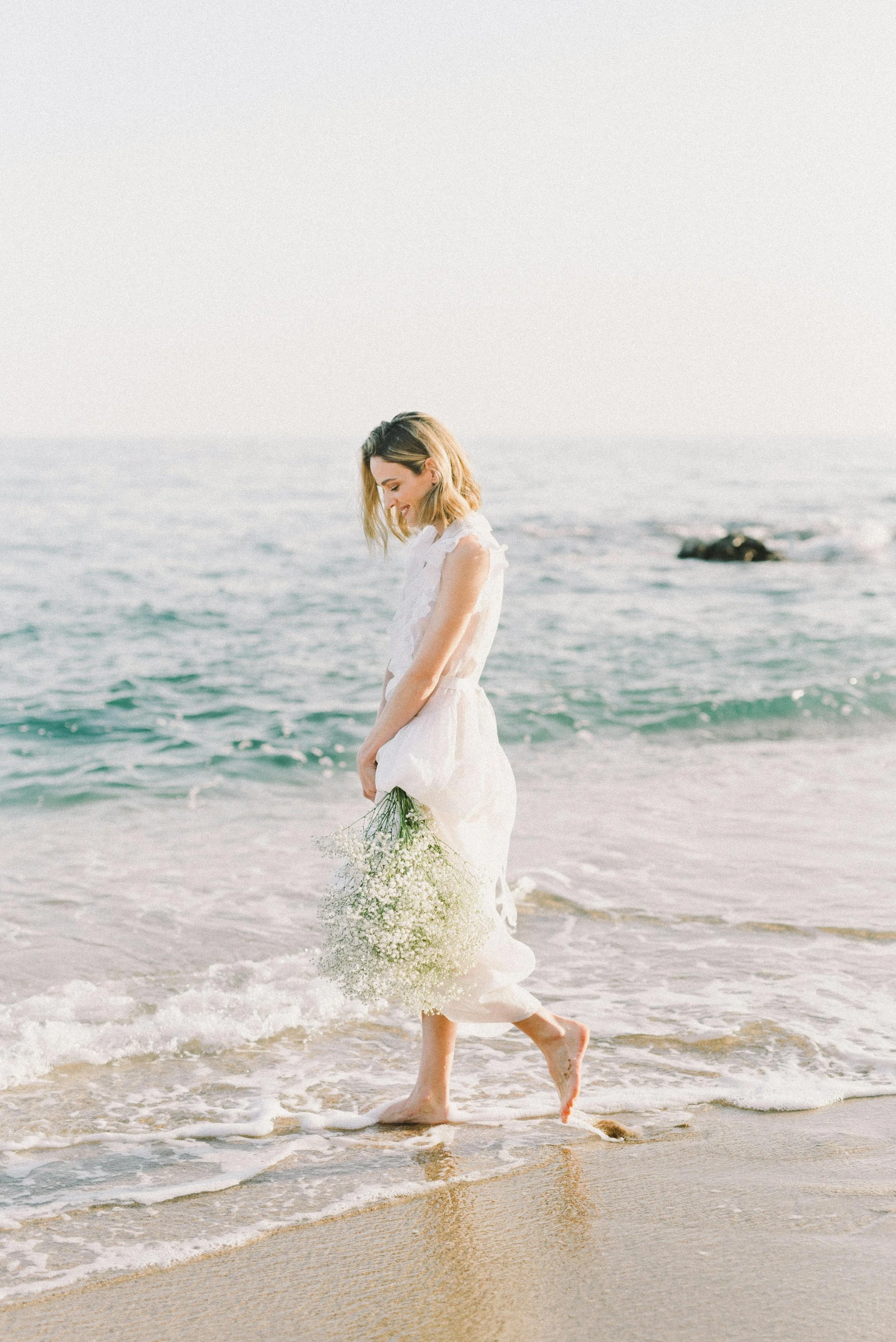 A woman in a white dress standing barefoot on the beach, holding a bouquet of white flowers, with the ocean in the background.