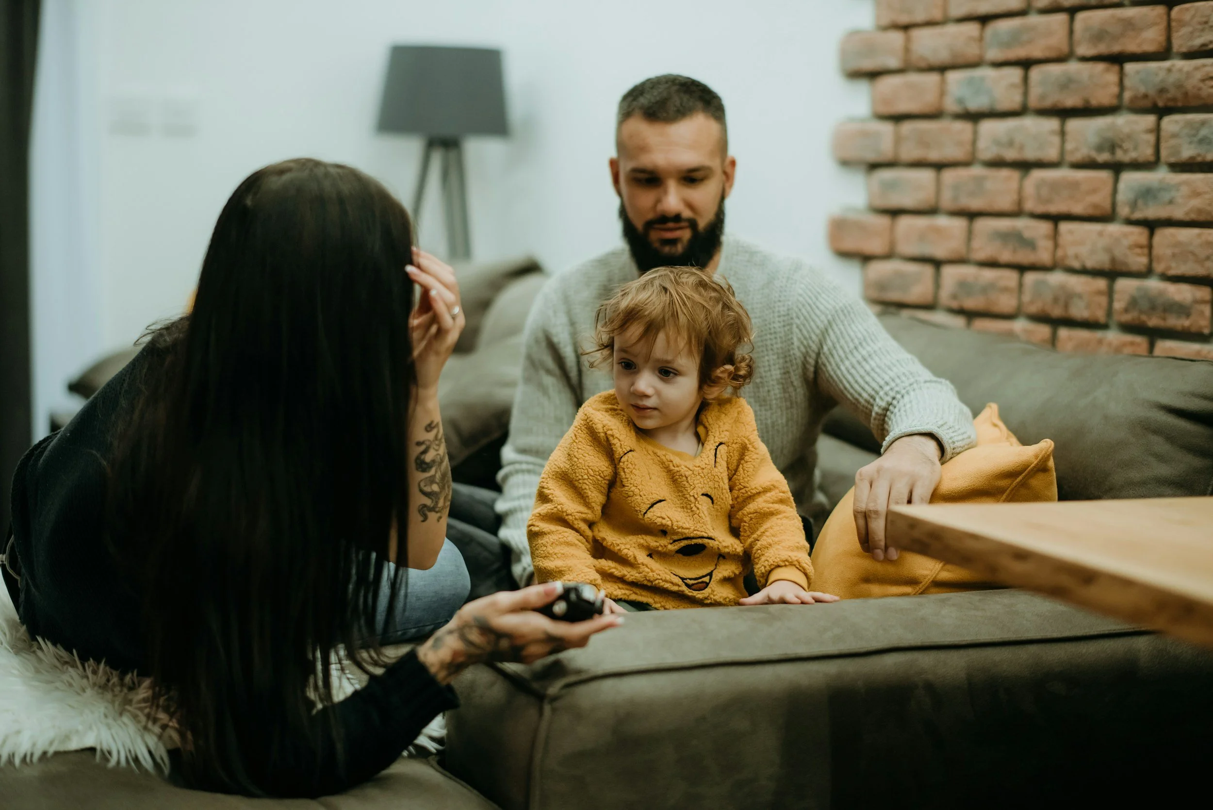 A woman with long dark hair and tattoos on her arm is talking to a man and a small child sitting on a couch in a living room. The woman covers part of her face with her hand. The man has a beard and is wearing a light gray sweater, and the child wears a yellow bear-themed sweater.