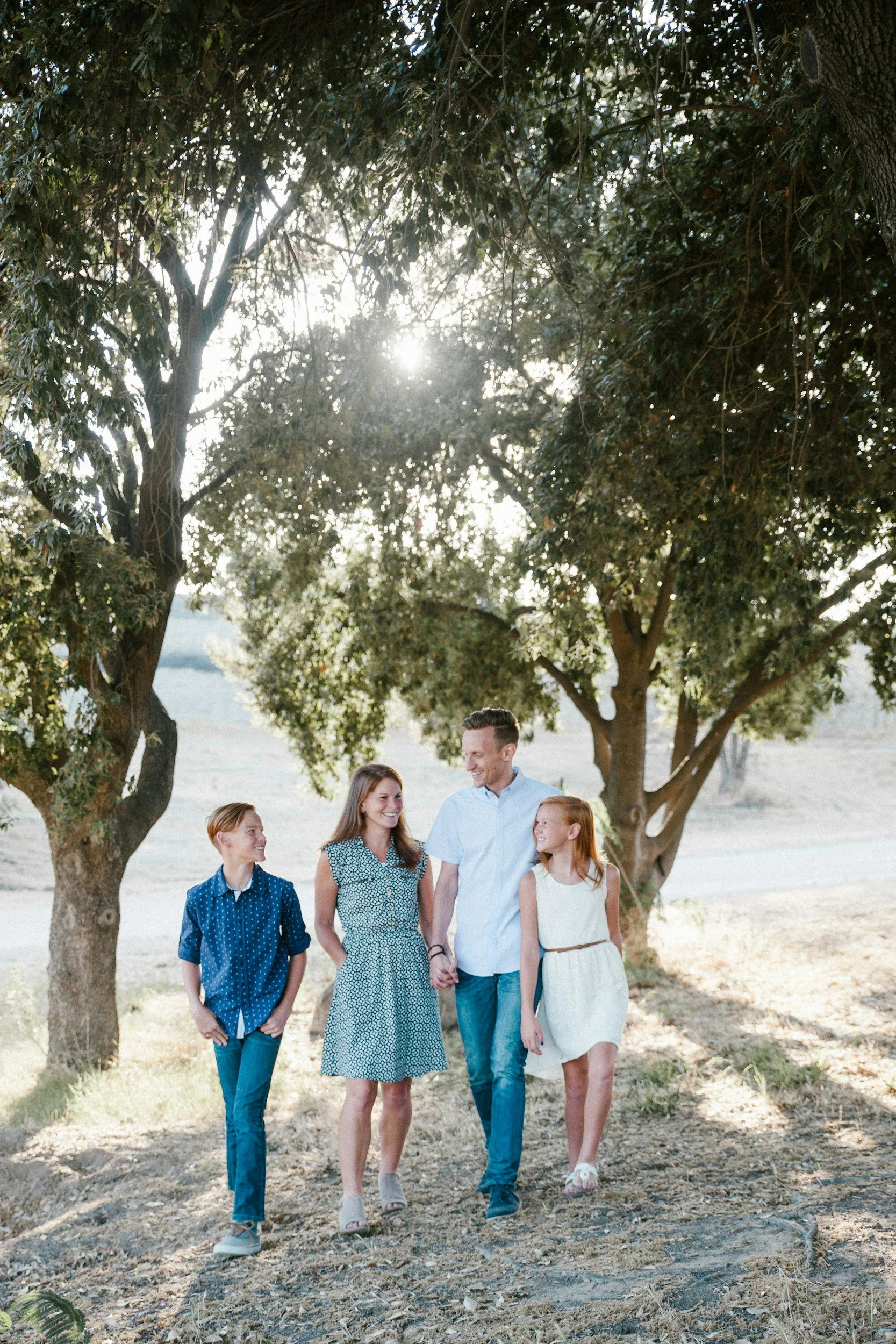 A family of four, two children and two adults, walking together outdoors on a sunny day under large trees.