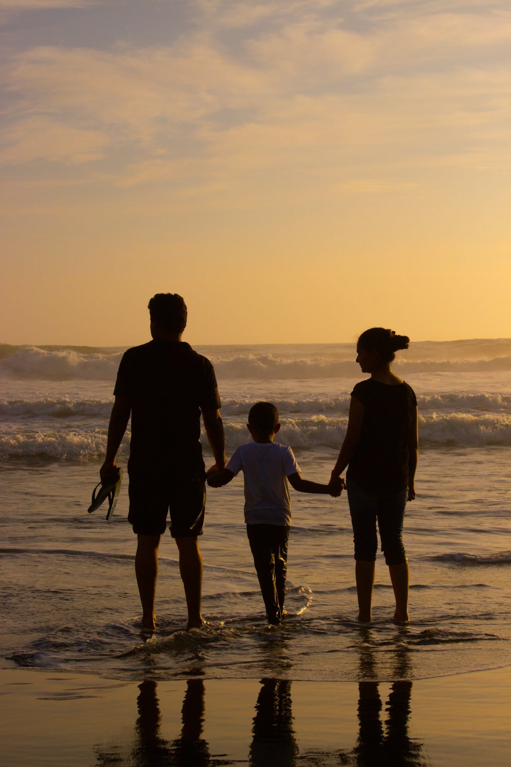 A family of three stands hand in hand at the beach during sunset, facing the ocean waves.