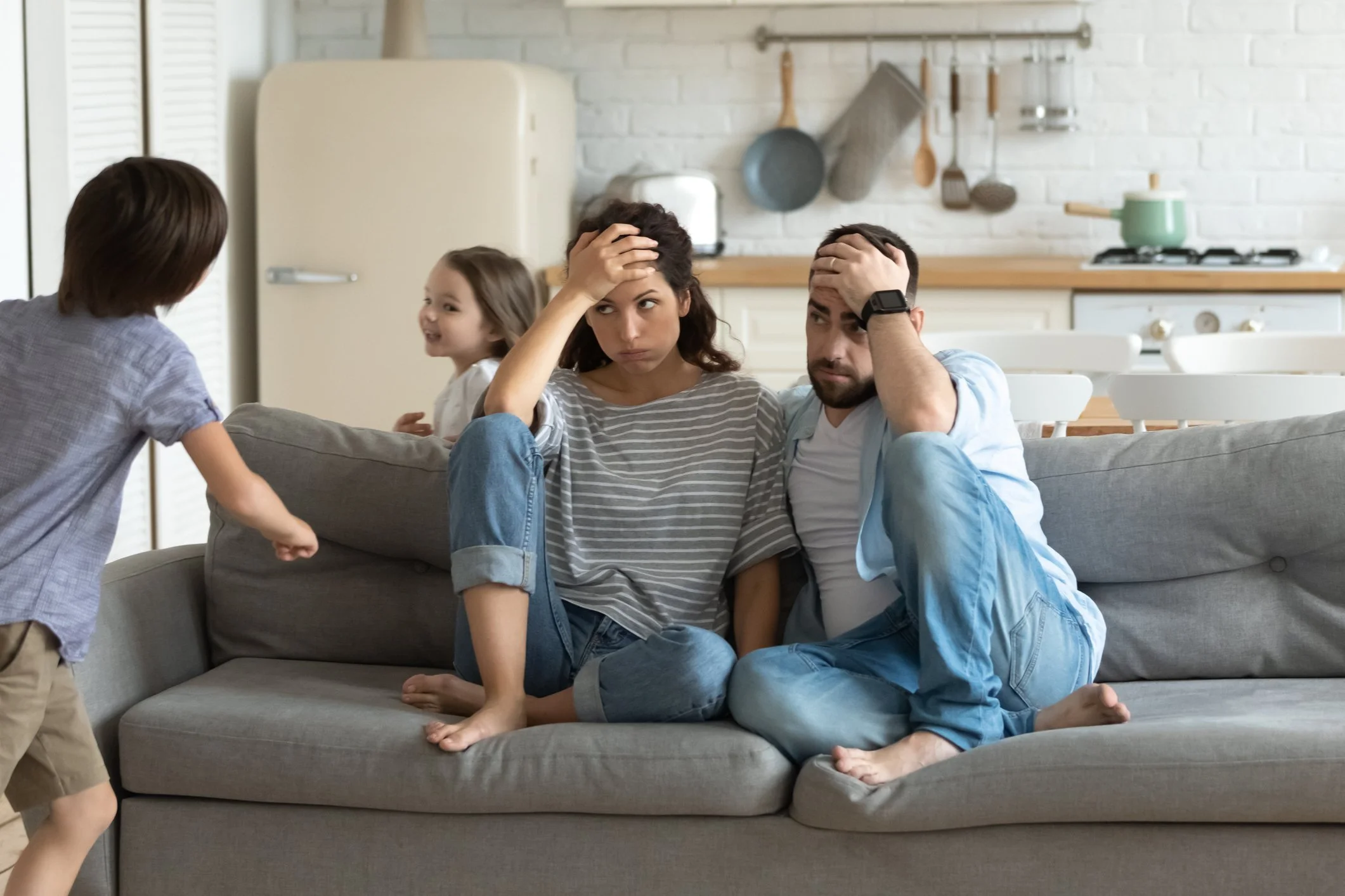 A family sitting on a gray couch with stressed expressions, children approaching them, in a kitchen background.