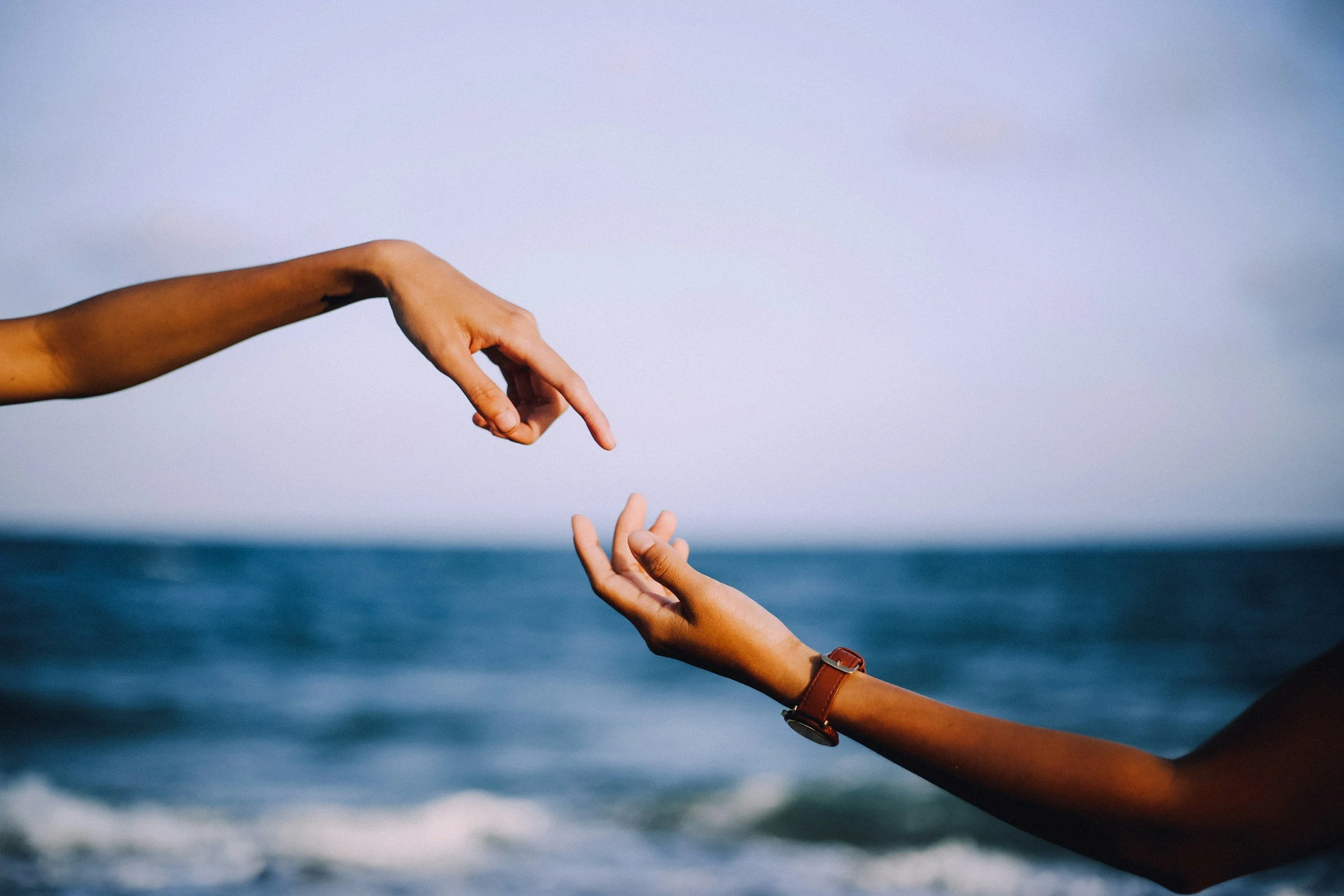 Two hands reaching towards each other above the ocean with a cloudy sky in the background.