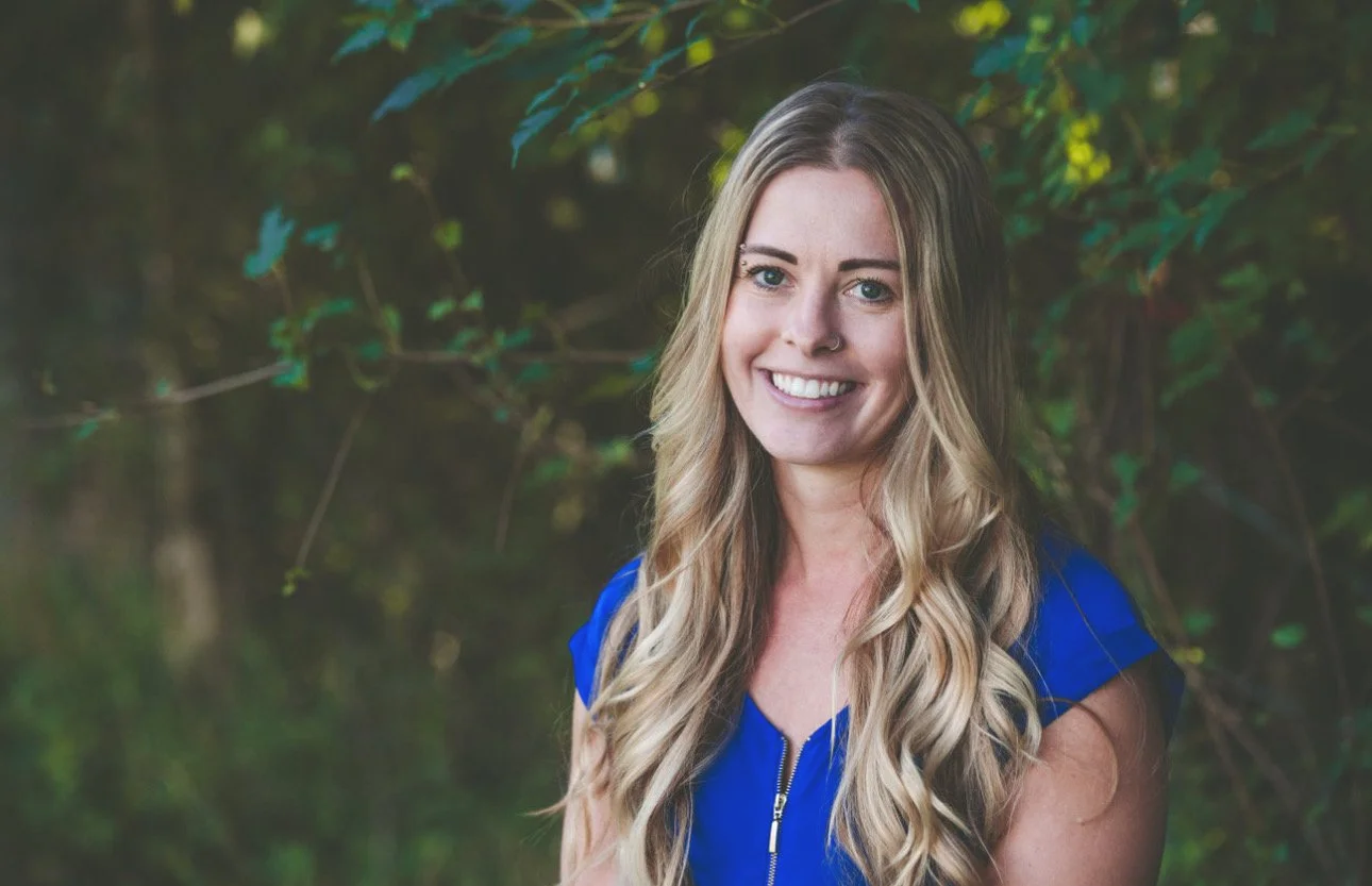 A young woman with long, wavy blonde hair and a nose piercing, smiling outdoors in front of greenery, wearing a royal blue top.
