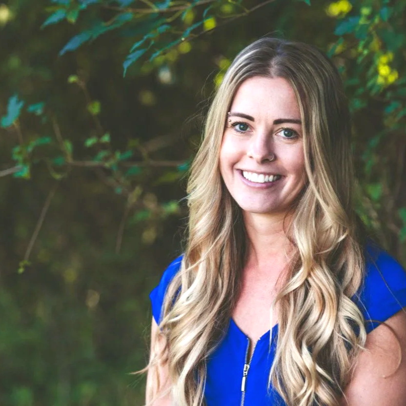 Trauma Therapist in Hopkins, MN with long blonde hair, blue eyes, and a nose piercing, wearing a blue top, standing outdoors with green foliage in the background.