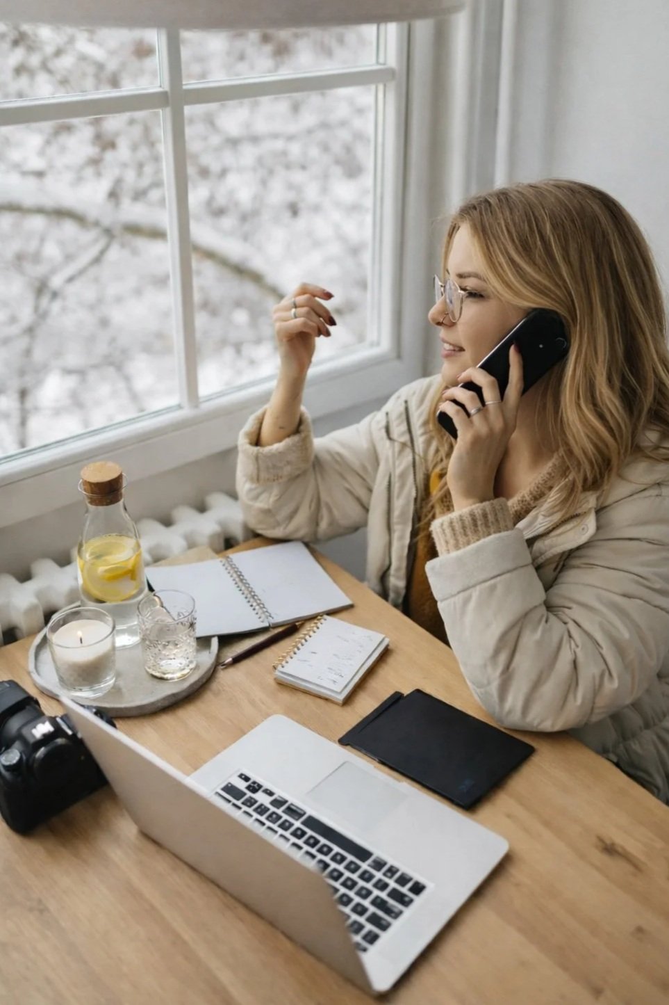 A woman sitting at a wooden desk near a window, talking on a landline phone, with a laptop, notebooks, a camera, candles, a water pitcher with lemon, and a glass on the desk, in Hopkins, Minnesota