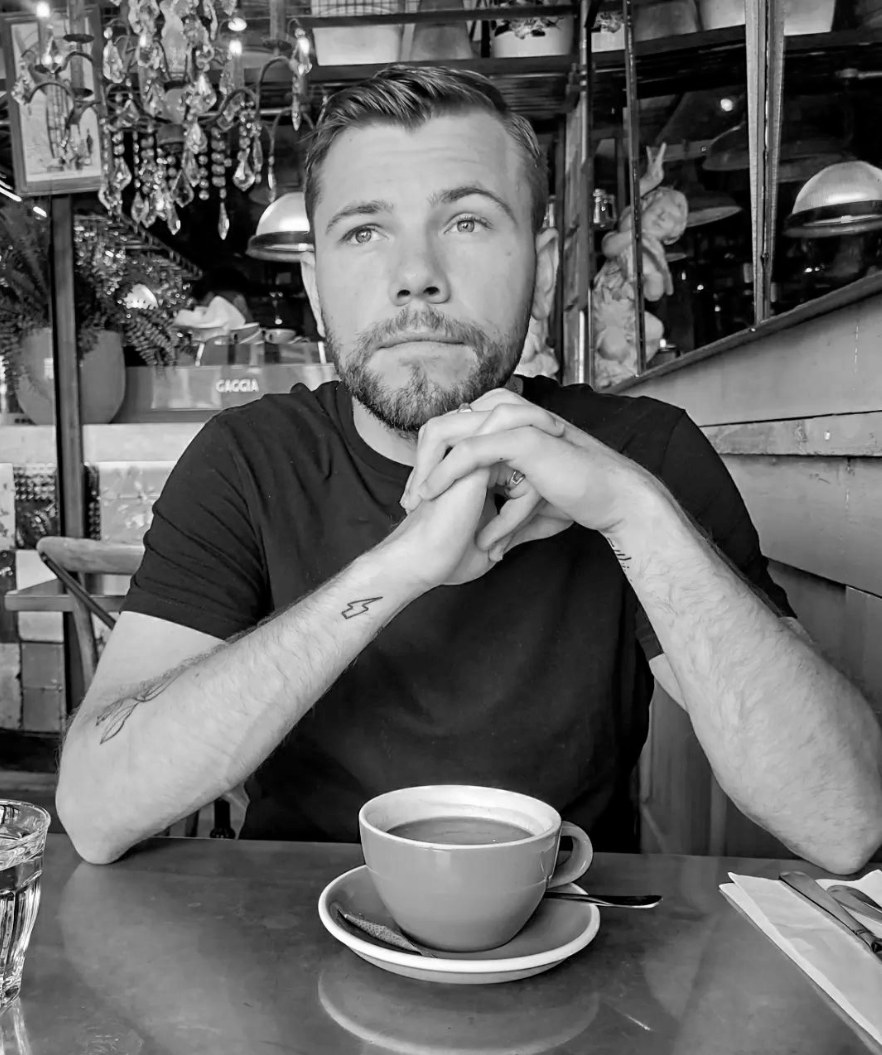 A man with a beard and short hair sitting at a table in a restaurant or cafe, with a cup of coffee in front of him and hands clasped under his chin, looking contemplative. The background has decorative elements and shelves.