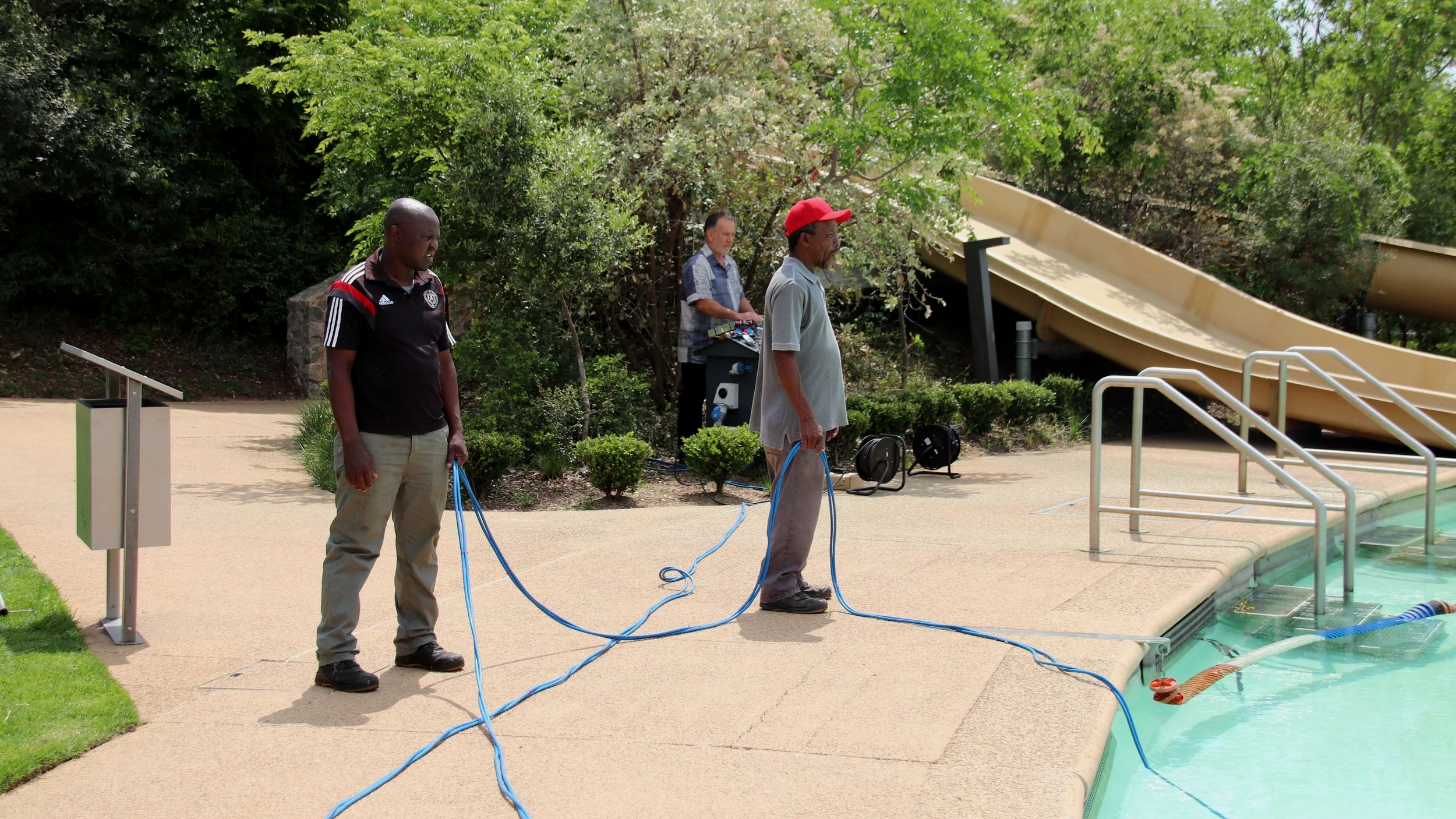 Three men standing beside a swimming pool holding a blue hose, with a waterslide and greenery in the background.