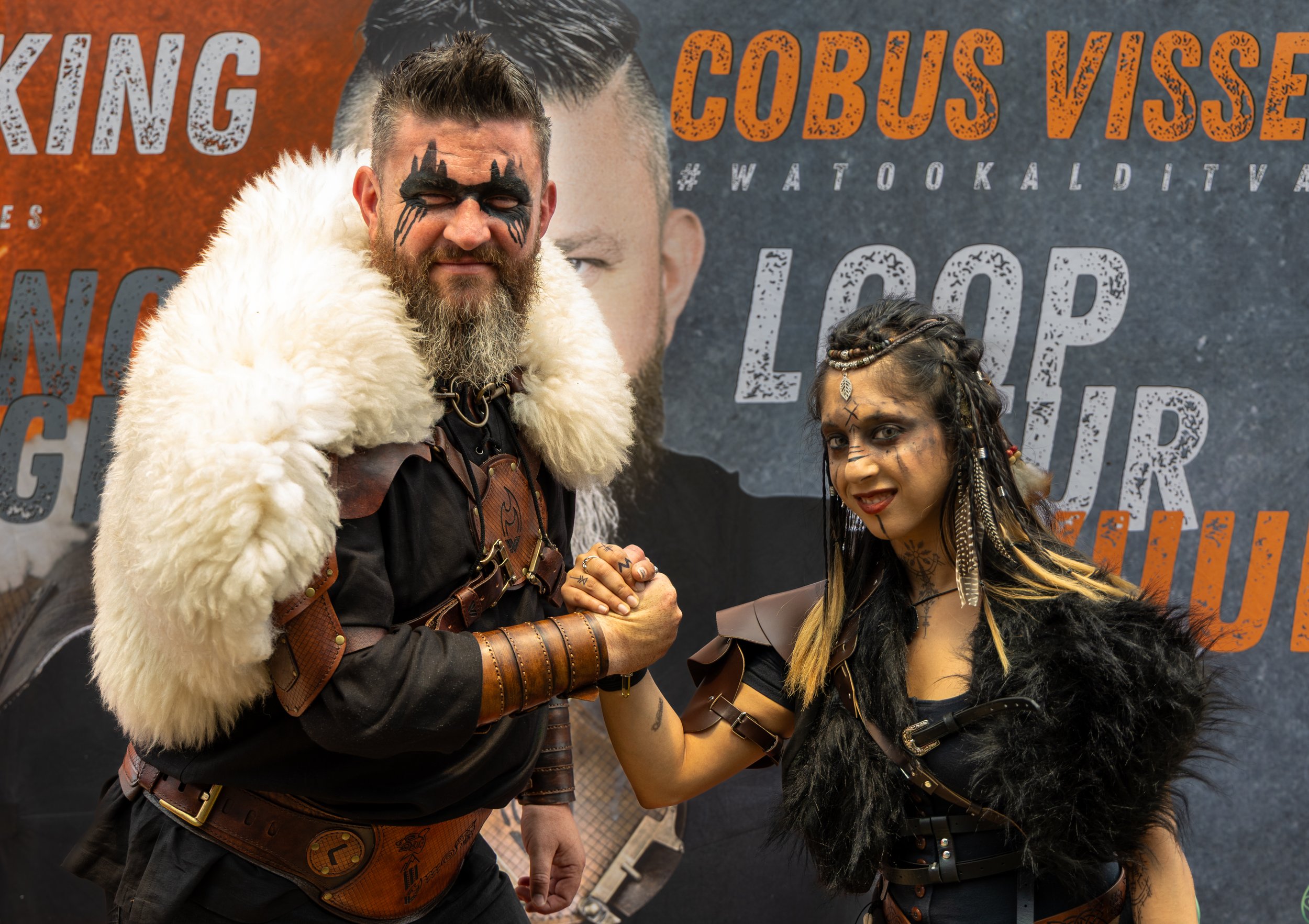 Two people dressed in warrior costumes pose in front of a promotional poster. The man on the left has face paint resembling a wolf, a thick beard, and wears a fur cloak over medieval armor. The woman on the right has braided hair, face tattoos, and wears a black costume with fur and leather accents. They are clasping hands in a fist bump, smiling at the camera.