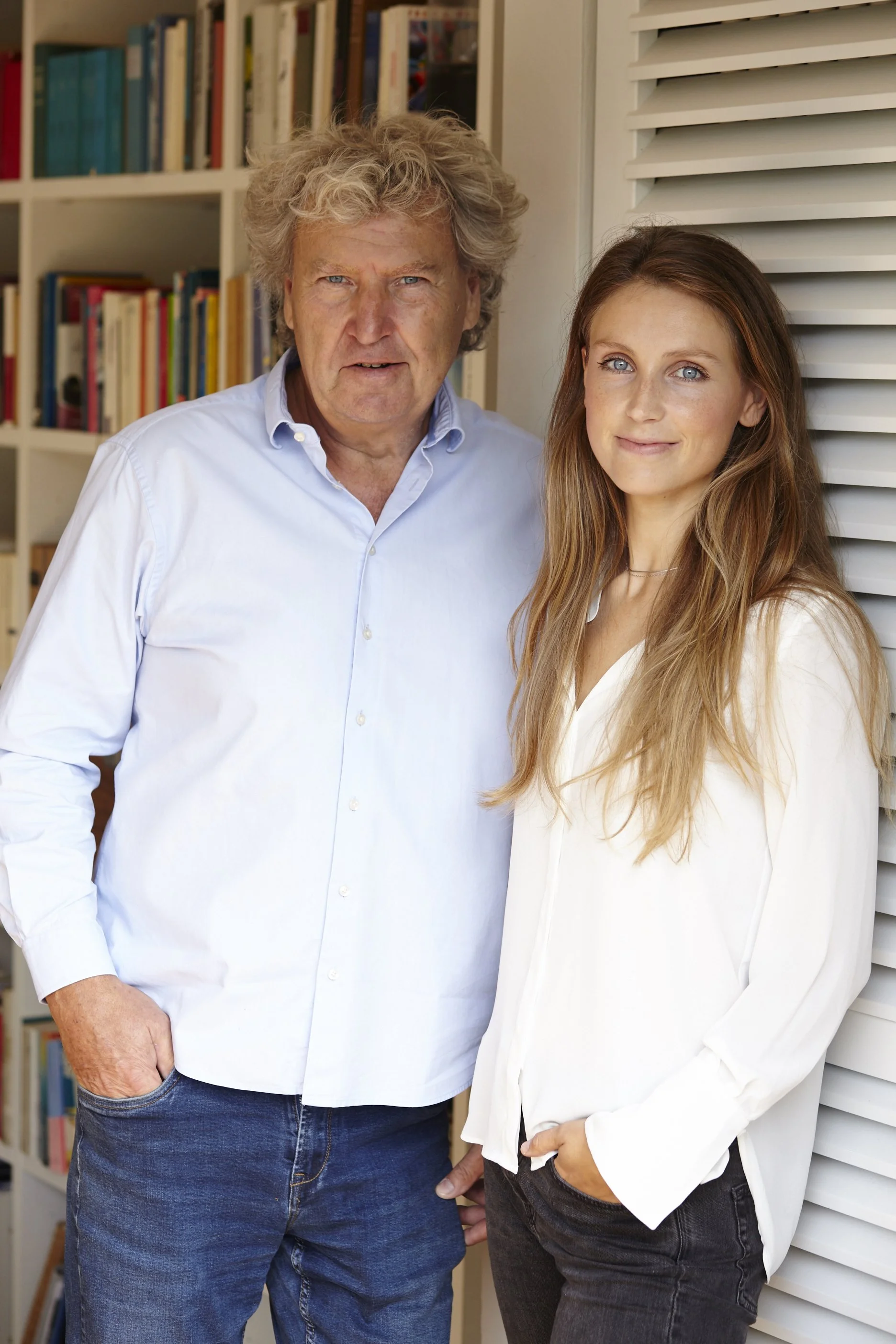 Ein älterer Mann und eine junge Frau stehen nebeneinander vor einer Bücherwand und einem weißen Lattendach. Sie sind beide leger gekleidet, der Mann trägt ein weißes Hemd und Jeans, die junge Frau trägt eine weiße Bluse und dunkle Jeans. Beide schauen in die Kamera.