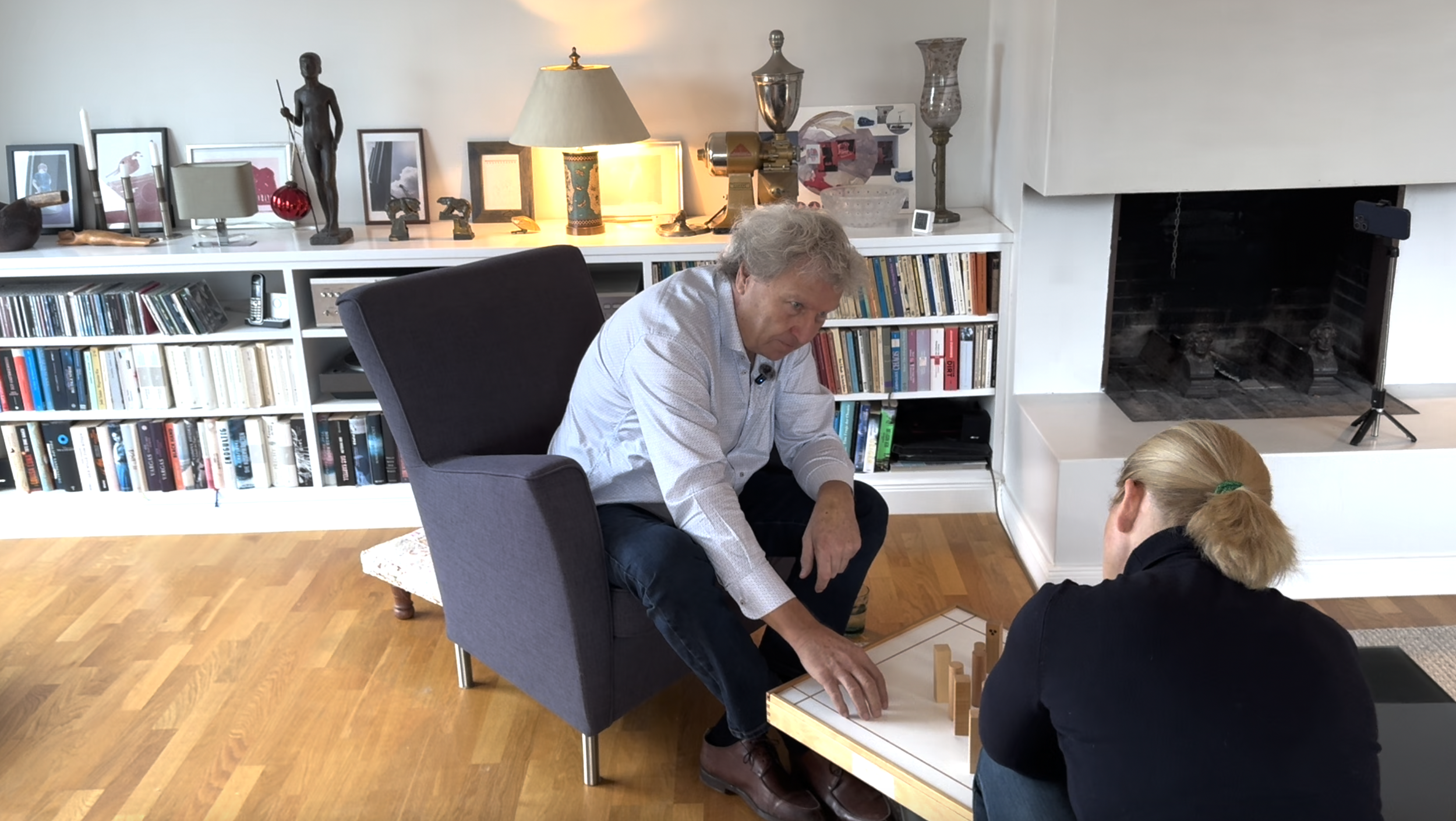Zwei Menschen am Familienbrett in einem Wohnzimmer, im Hintergrund sind Bücherregale, Lampen, Kunstgegenstände und Dekorationen sichtbar.