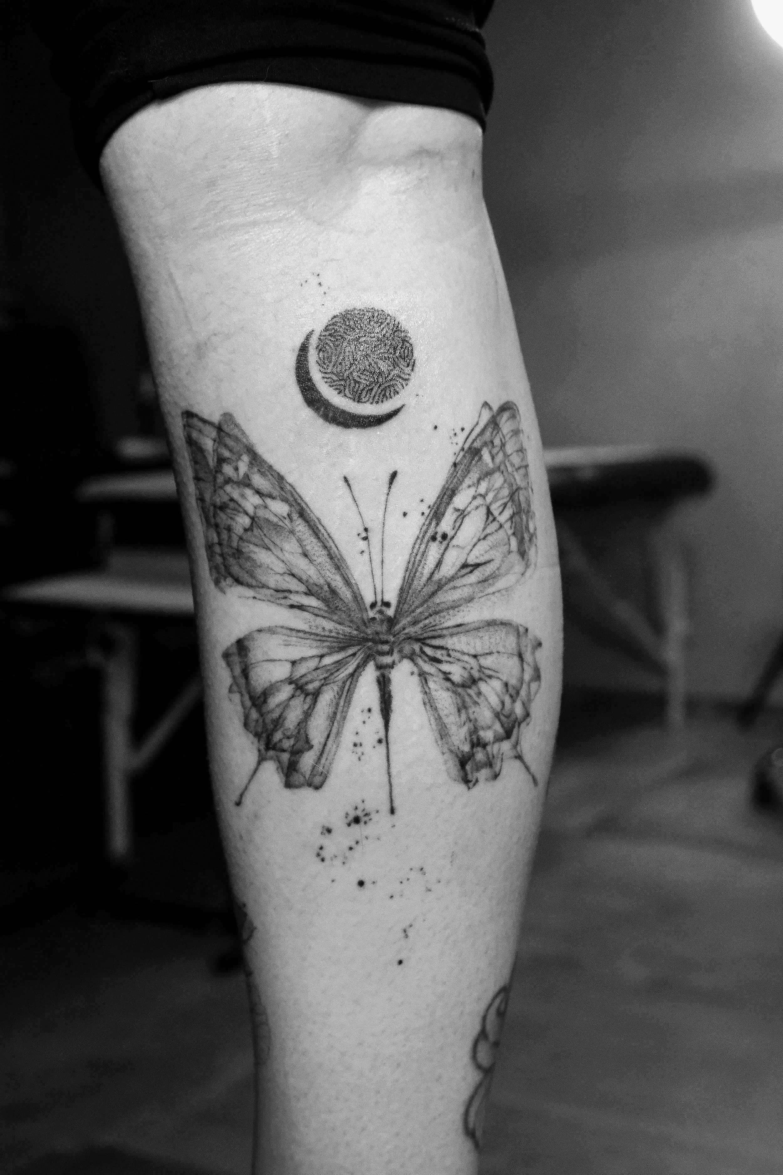 Close-up of a person's arm with an x-ray style, very delicate butterfly fineline tattoo and a small moon and geometric design tattoo above it, black and white photo.