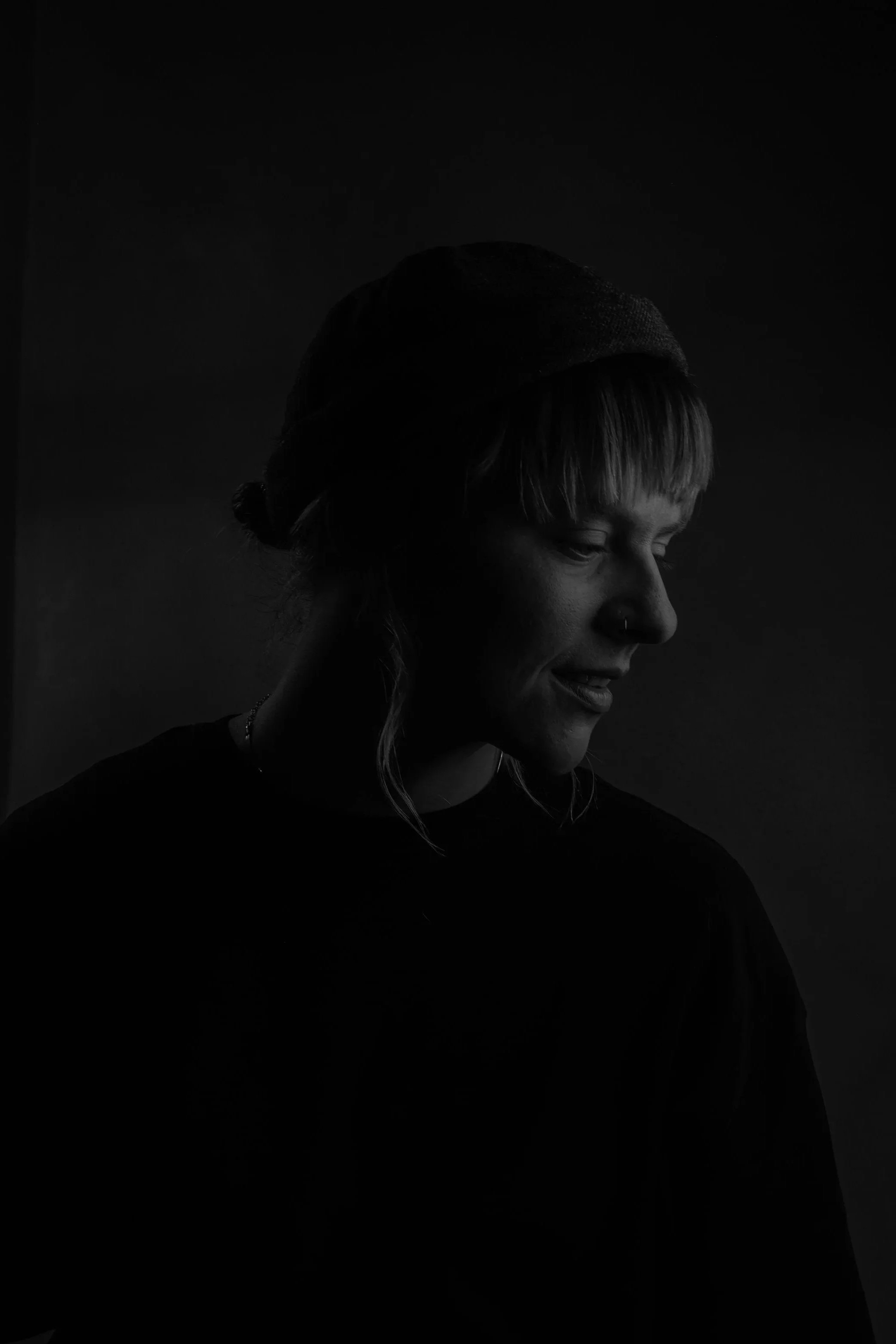 Portrait picture of Ola Getka, tattoo artist and choreographer based in Iceland, wearing a dark beanie and a nose ring, smiling and looking down, against a dark background.
